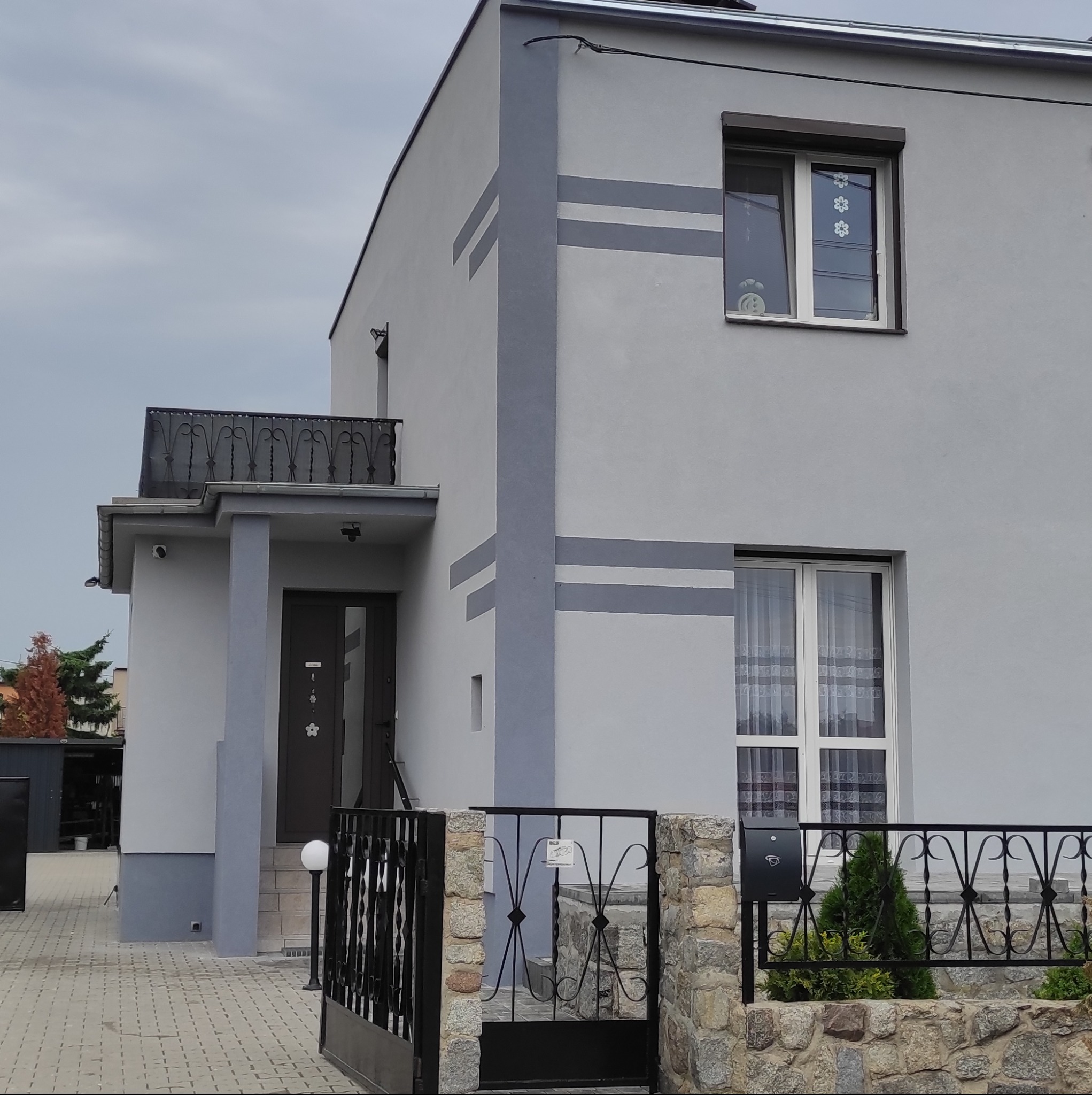 Dwukondygnacyjny dom jednorodzinny z szarą elewacją, ozdobiony poziomymi pasami w ciemniejszym odcieniu szarości, widoczny balkon z kutą balustradą, okna z białymi ramami i firankami,...