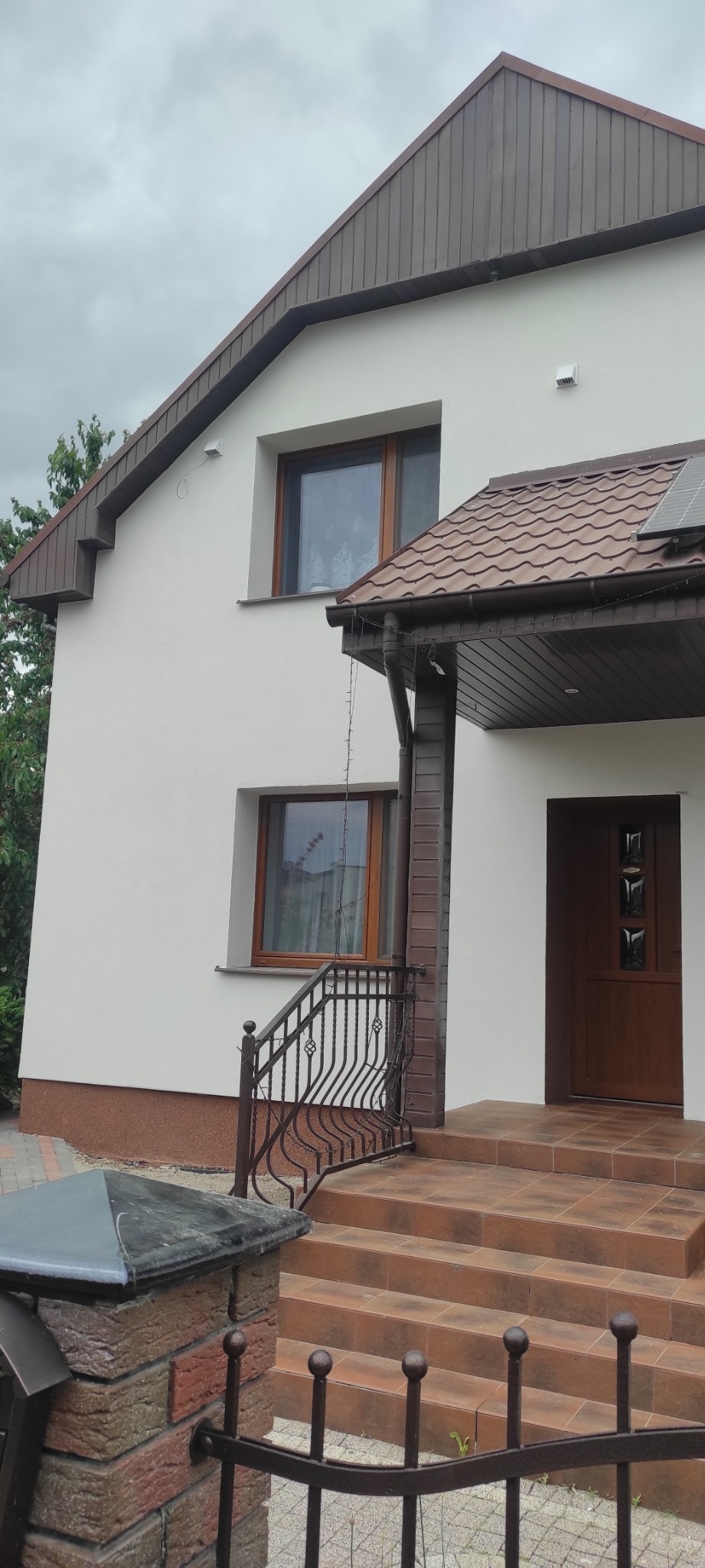 Wykończony dom jednorodzinny z białą elewacją, brązowym dachem z blachodachówki i drewnianymi oknami, widoczne schody wejściowe wyłożone brązowymi płytkami i ozdobna metalowa balustrada.