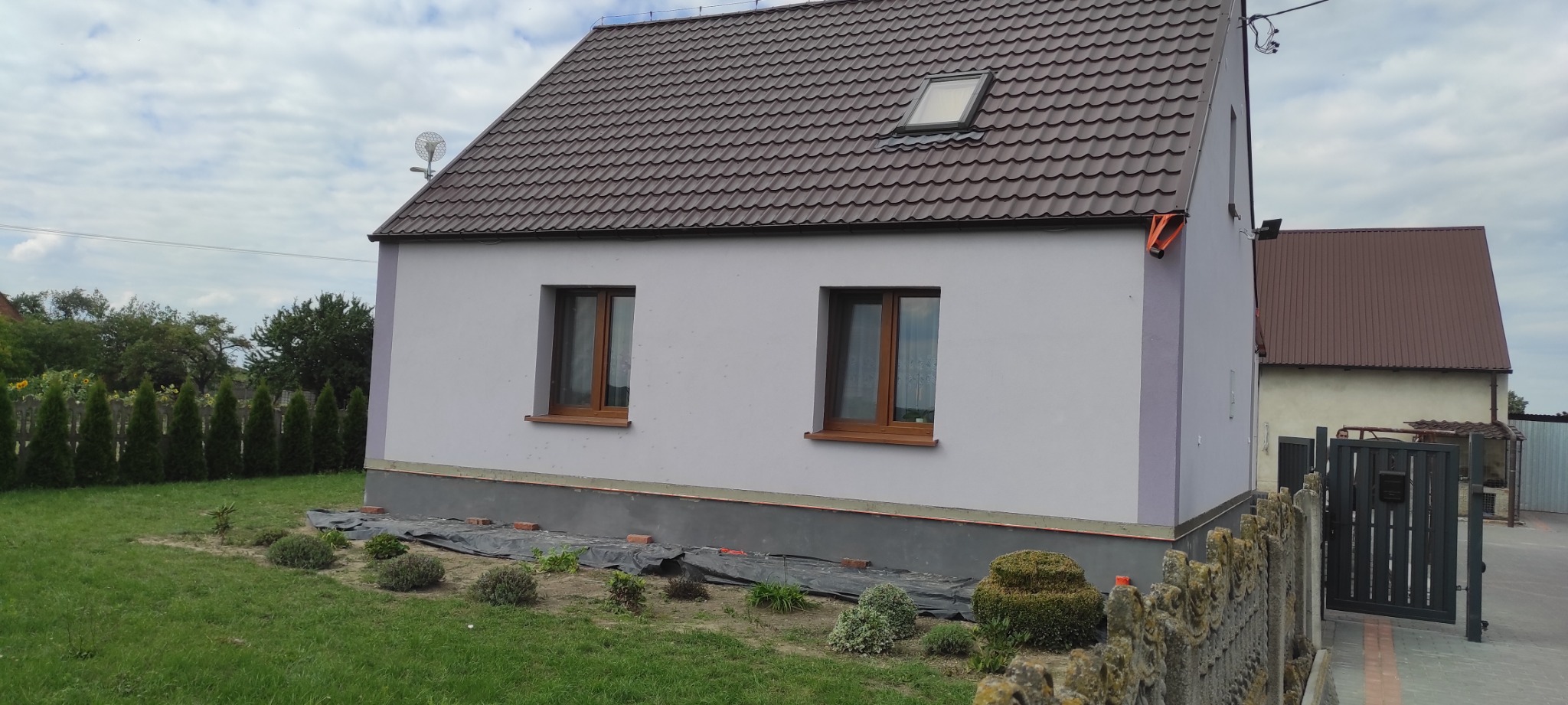 Dom jednorodzinny z nową, brązową blachodachówką i oknami w drewnianych ramach, świeżo pomalowany na jasny kolor, z widocznym fragmentem fundamentu zabezpieczonego folią.
