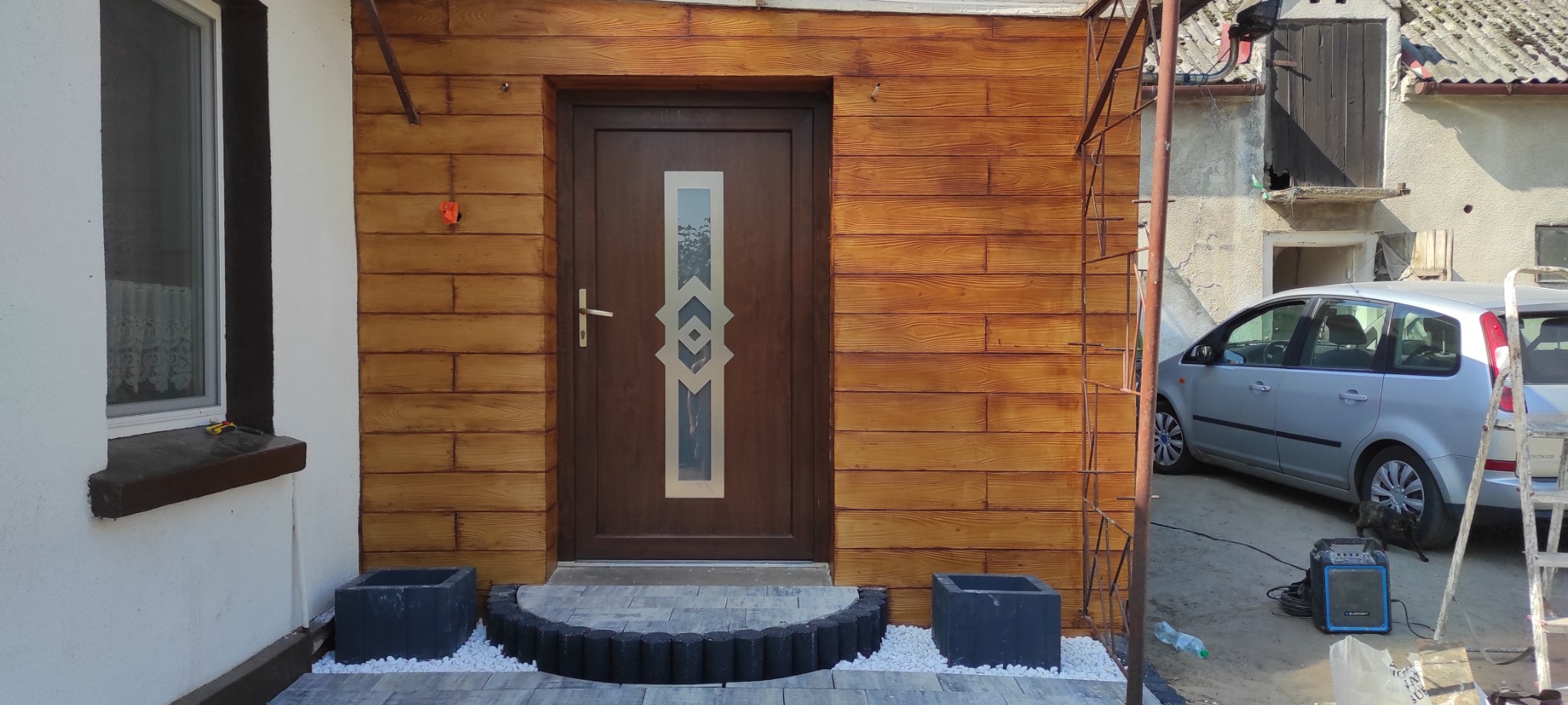 Elewacja budynku z drewnopodobną strukturą, brązowe drzwi z szybą o geometrycznym wzorze, obok okno z brązowymi ramami, przed wejściem dwa czarne kwadratowe kwietniki z białymi kamykami i półokrąg...