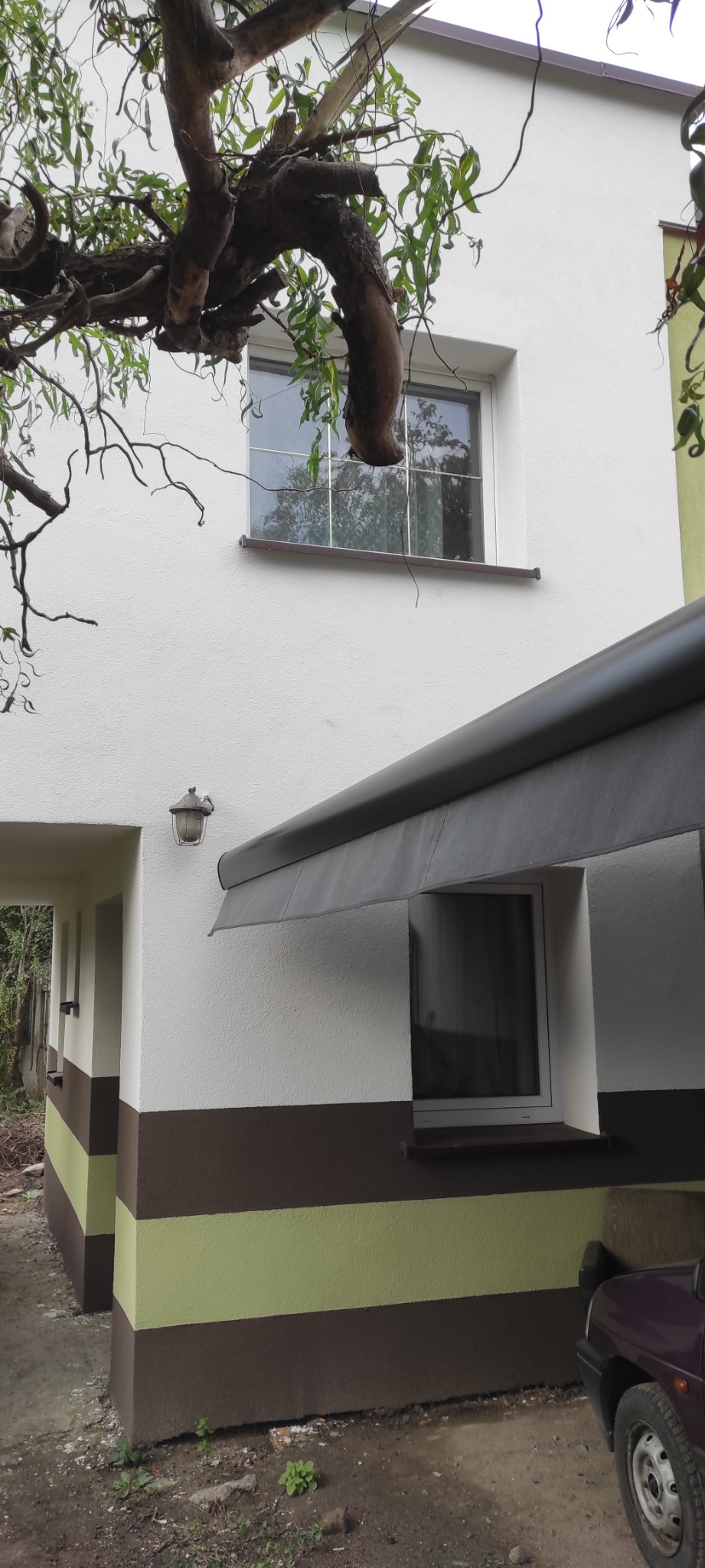Odnowiona elewacja budynku z dekoracyjnymi pasami w odcieniach brązu i zieleni, widoczny fragment samochodu i gałąź drzewa zwisająca nad oknem na piętrze.