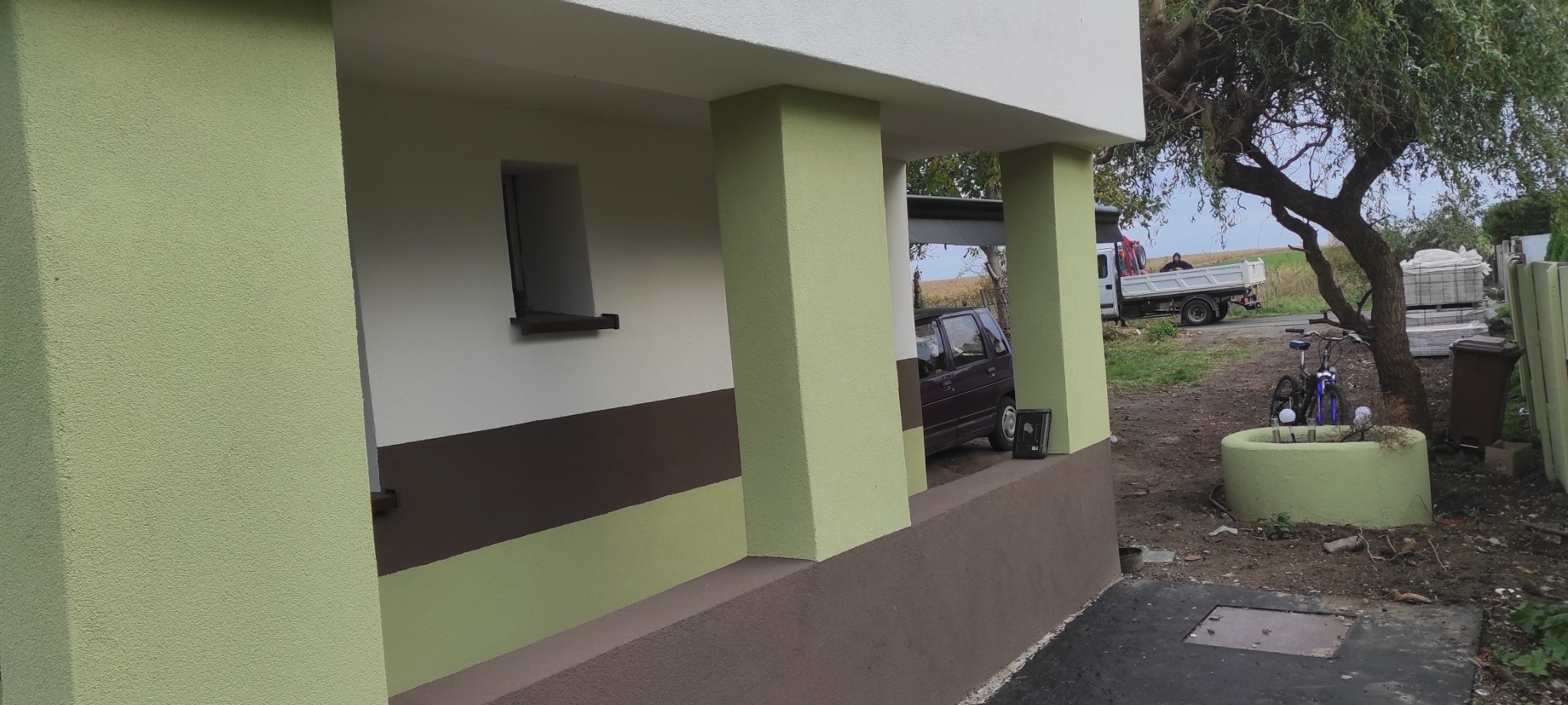 Świeżo pomalowana elewacja domu z kolumnami w odcieniach zieleni i brązu, widoczne okno, zaparkowany samochód i ciężarówka w tle.