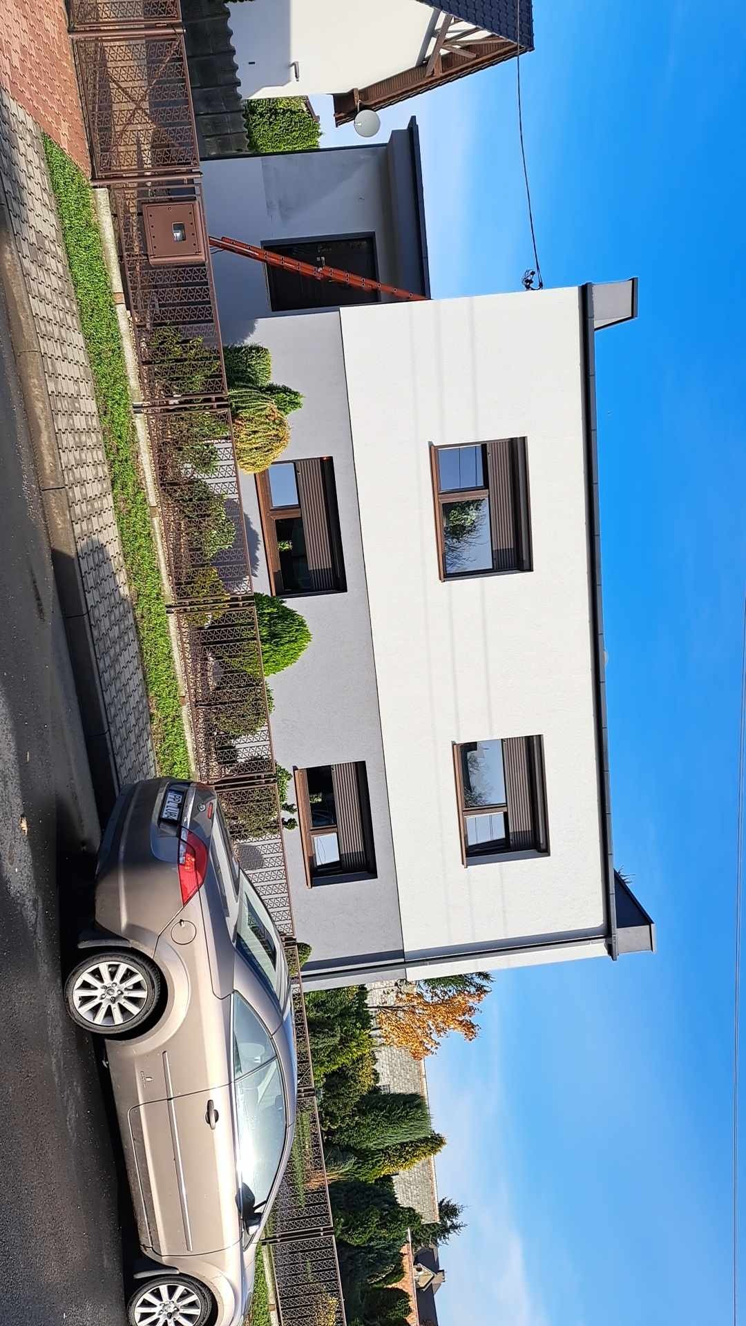 Nowoczesny, dwukondygnacyjny dom z elewacją w kolorze szarości i bieli, z brązowymi oknami i metalowym ogrodzeniem, widziany z perspektywy ulicy, z zaparkowanym samochodem w kolorze beżowym.