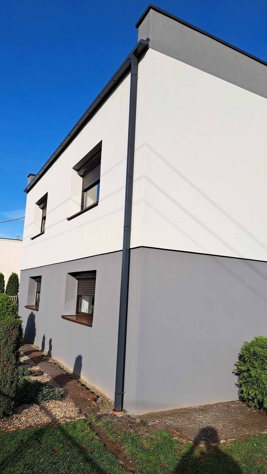 Dwukondygnacyjny dom z nową, dwukolorową elewacją: biała góra i szary dół. Widoczne okna z brązowymi ramami i szarymi roletami oraz ciemna rynna.