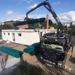 Czarny dźwig Kesla 2124L podnosi wiązkę zielonych łat dachowych nad budynkiem z białych bloczk&oacute;w betonowych na placu budowy w słoneczny dzień.