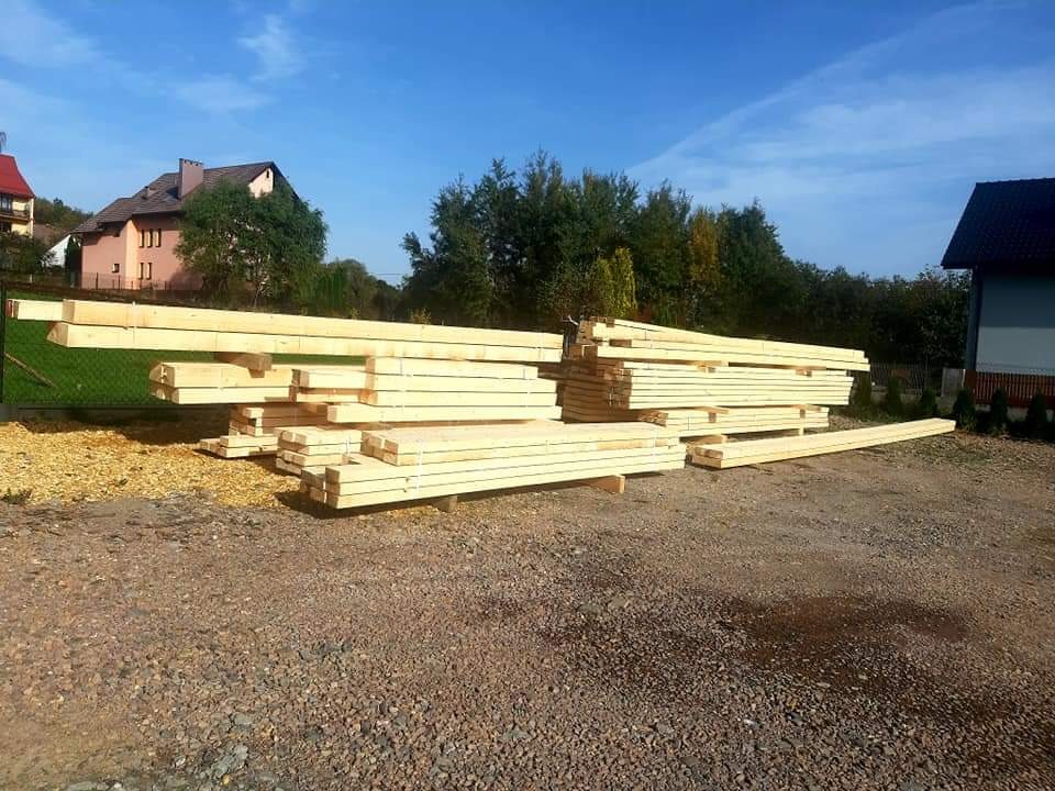 Stosy świeżo przetartego, jasnego drewna ułożone na placu budowy, widoczne budynki mieszkalne w tle pod błękitnym niebem.