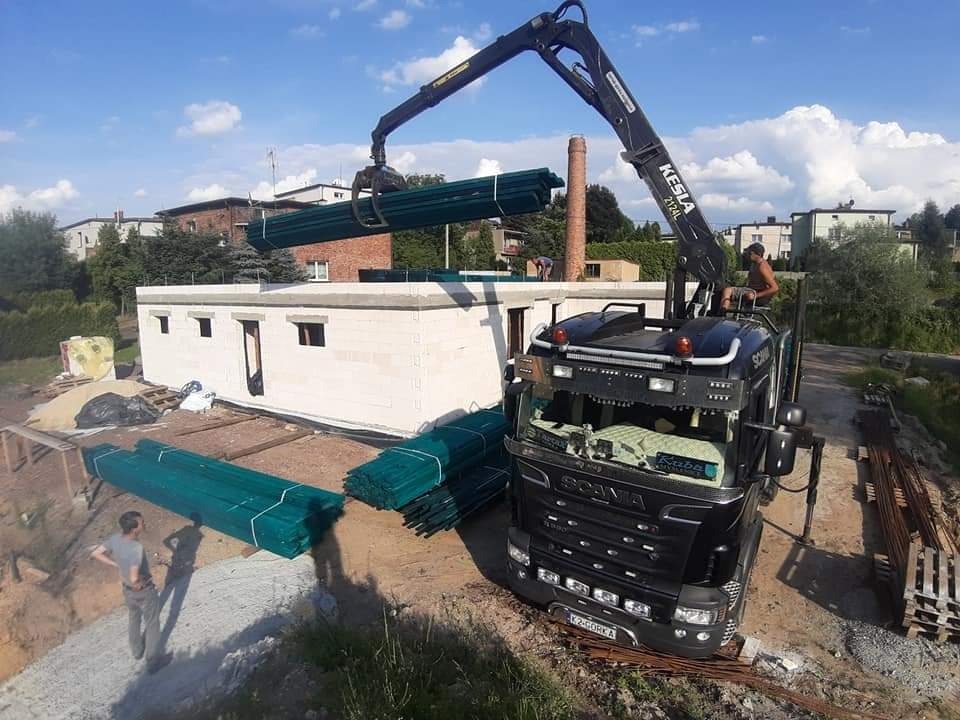 Czarny dźwig Kesla 2124L podnosi wiązkę zielonych łat dachowych nad budynkiem z białych bloczków betonowych na placu budowy w słoneczny dzień.