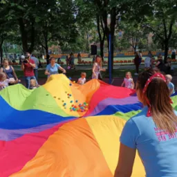 Kolorowa chusta animacyjna z małymi piłeczkami w centrum, wokół której stoją dzieci i dorośli podczas zabawy na świeżym powietrzu.