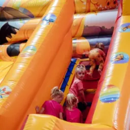 Dzieci wspinają się na dmuchaną zjeżdżalnię z motywami zwierzęcymi i tęczowymi naklejkami, jedna dziewczynka z pomalowaną twarzą patrzy w obiektyw.