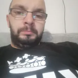 Portret mężczyzny w okularach i czarnej koszulce z białym nadrukiem korony, z widocznymi kablami na ścianie w tle.