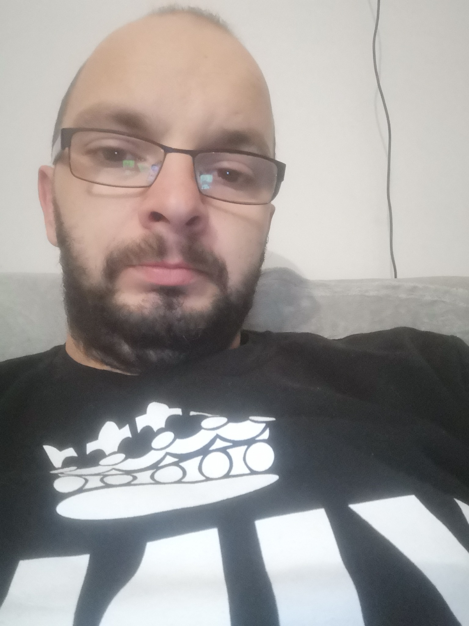 Portret mężczyzny w okularach i czarnej koszulce z białym nadrukiem korony, z widocznymi kablami na ścianie w tle.