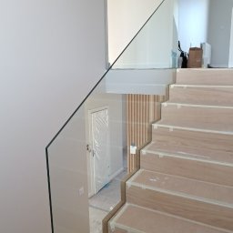 DeGlass- usługi szklarskie - Szklana balustrada zamontowana na drewnianych schodach w trakcie wykańczania; widoczna folia zabezpieczająca na drzwiach w tle.