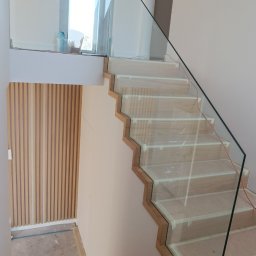 DeGlass- usługi szklarskie - Szklana balustrada schodowa z drewnianymi stopniami w trakcie montażu, zabezpieczona taśmą malarską, widok z góry na dół schodów.
