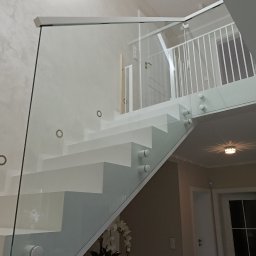 DeGlass- usługi szklarskie - Nowoczesne, białe schody z szklaną balustradą mocowaną punktowo, widok z dołu na górę, z elementami dekoracyjnymi w tle.