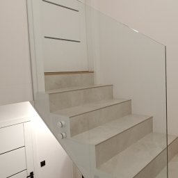 DeGlass- usługi szklarskie - Nowoczesne schody z jasnymi stopniami i szklaną balustradą mocowaną na okrągłych uchwytach, widok z dołu na białe drzwi z poziomymi frezowaniami.