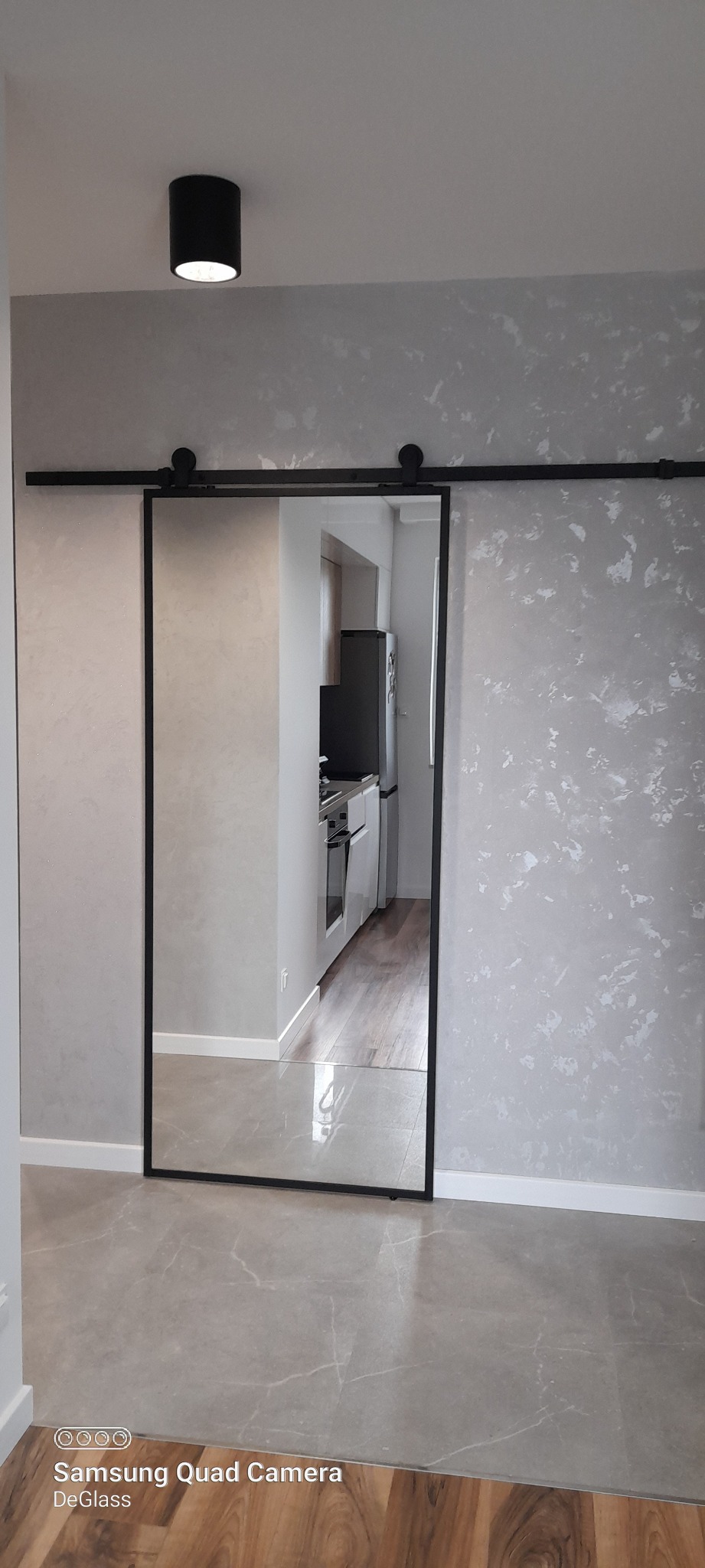 Przesuwne drzwi lustrzane z czarną ramą i systemem szynowym, widok na kuchnię w tle.