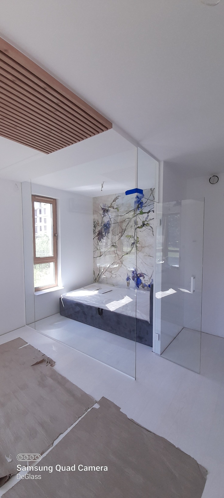 Szklana zabudowa w pokoju, widoczna szyba oddzielająca przestrzeń z łóżkiem i tapetą z motywem kwiatowym, częściowo zabezpieczona folią, na podłodze resztki folii ochronnej.