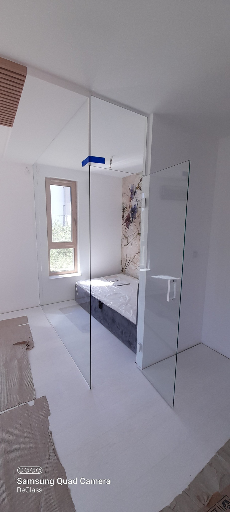 Montaż szklanej ścianki działowej w pokoju, widoczne zabezpieczenia na górnej krawędzi szkła, podłoga zabezpieczona kartonem, w tle łóżko i okno.