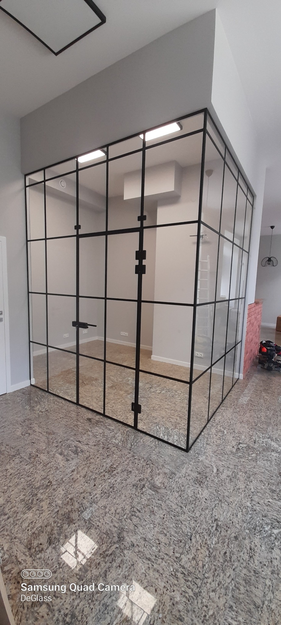 Nowoczesna szklana ścianka działowa z czarnymi profilami, tworząca przeszklone biuro w jasnym pomieszczeniu, widoczne drzwi z klamką.