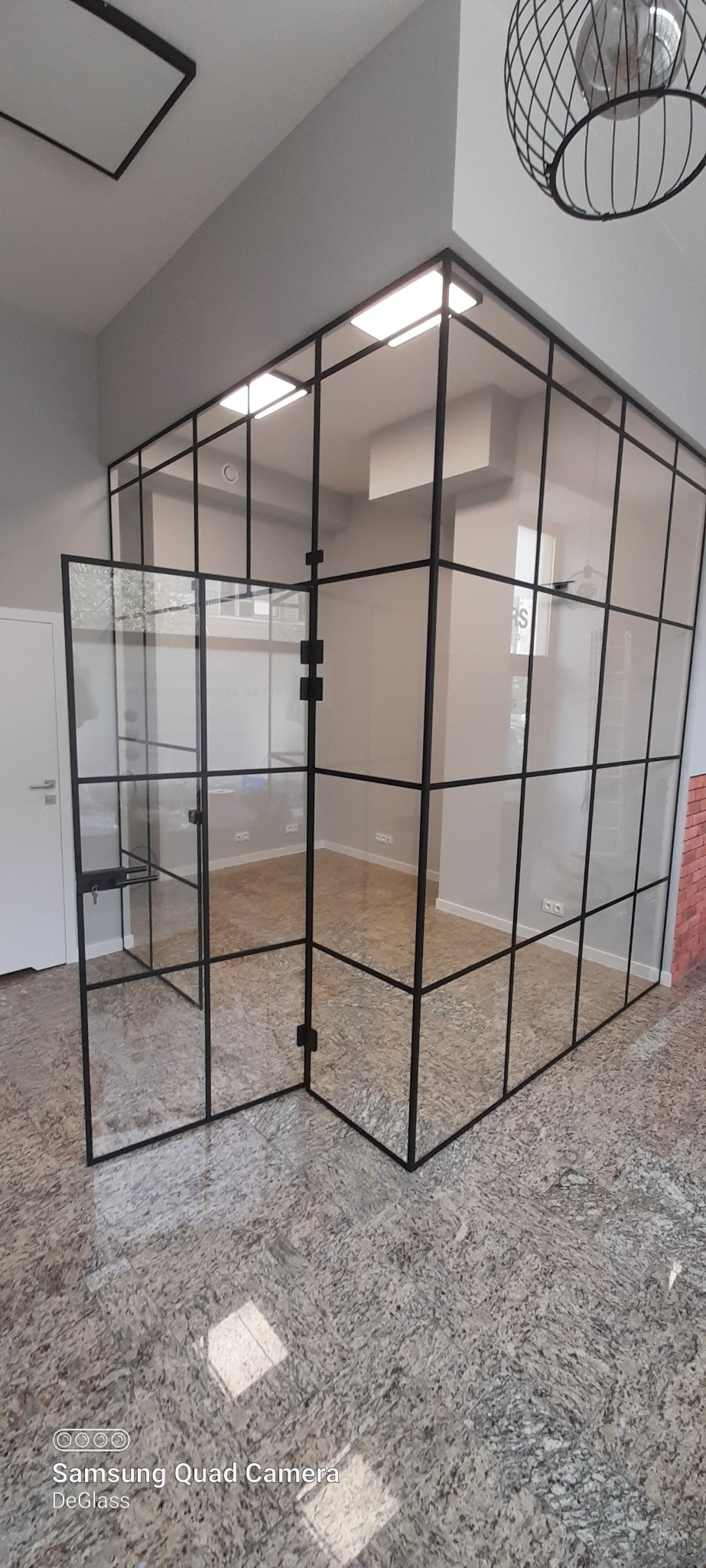 Nowoczesna szklana ścianka działowa z czarnymi profilami, częściowo otwarta, w minimalistycznym biurze z jasnymi ścianami i podłogą imitującą kamień.