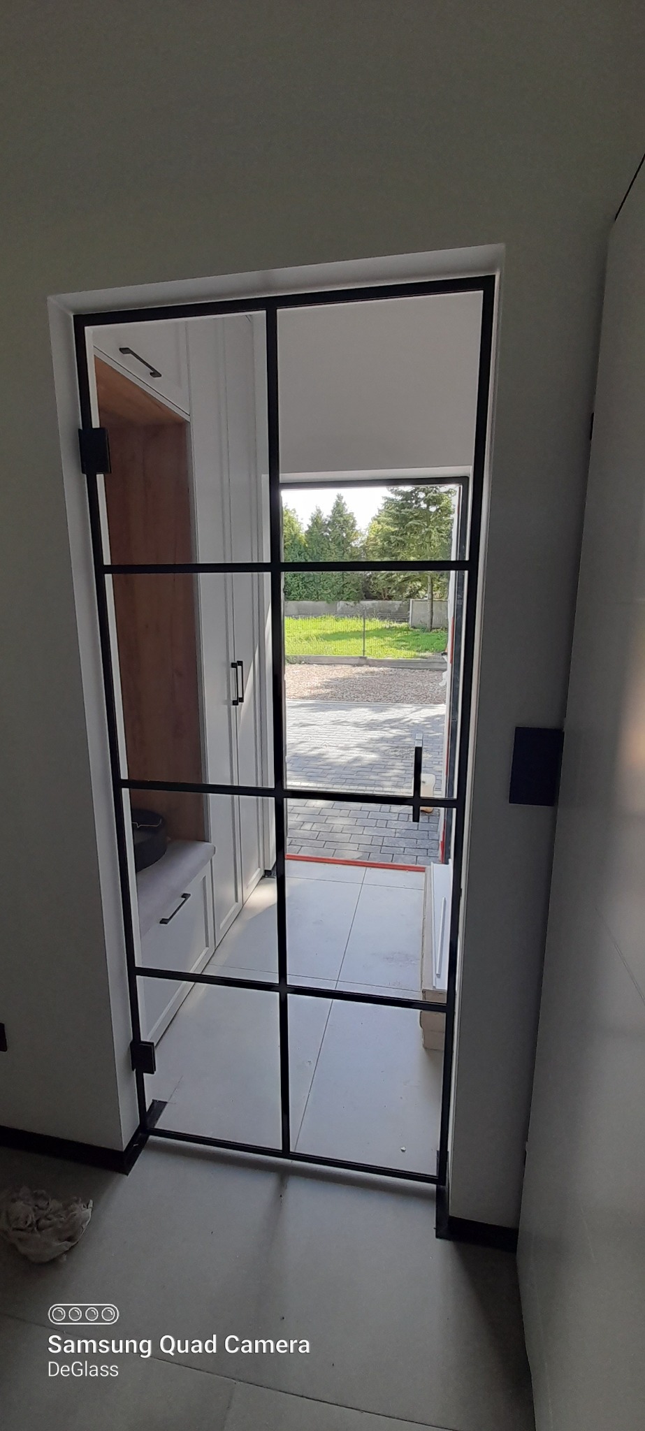 Wnętrze domu z widokiem na zewnątrz przez szklane drzwi z czarnymi szprosami, obok wnęka z zabudową meblową w kolorze białym.