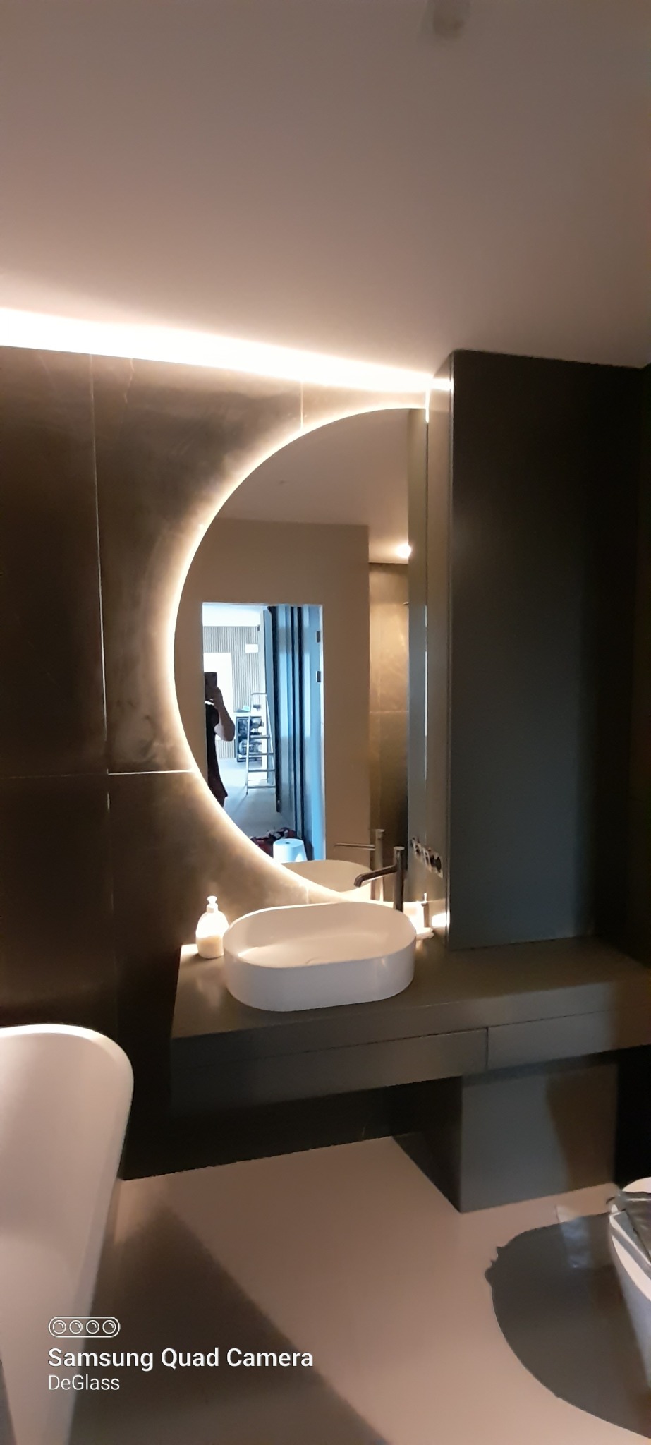 Nowoczesna łazienka z okrągłym lustrem z podświetleniem LED, umywalką nablatową i ciemnymi płytkami na ścianach.