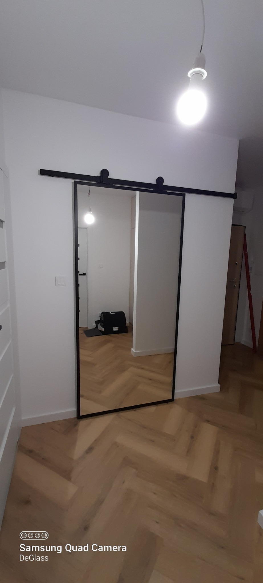 Przesuwne drzwi lustrzane z czarną ramą zawieszone na szynie, widoczne na tle parkietu ułożonego w jodełkę.