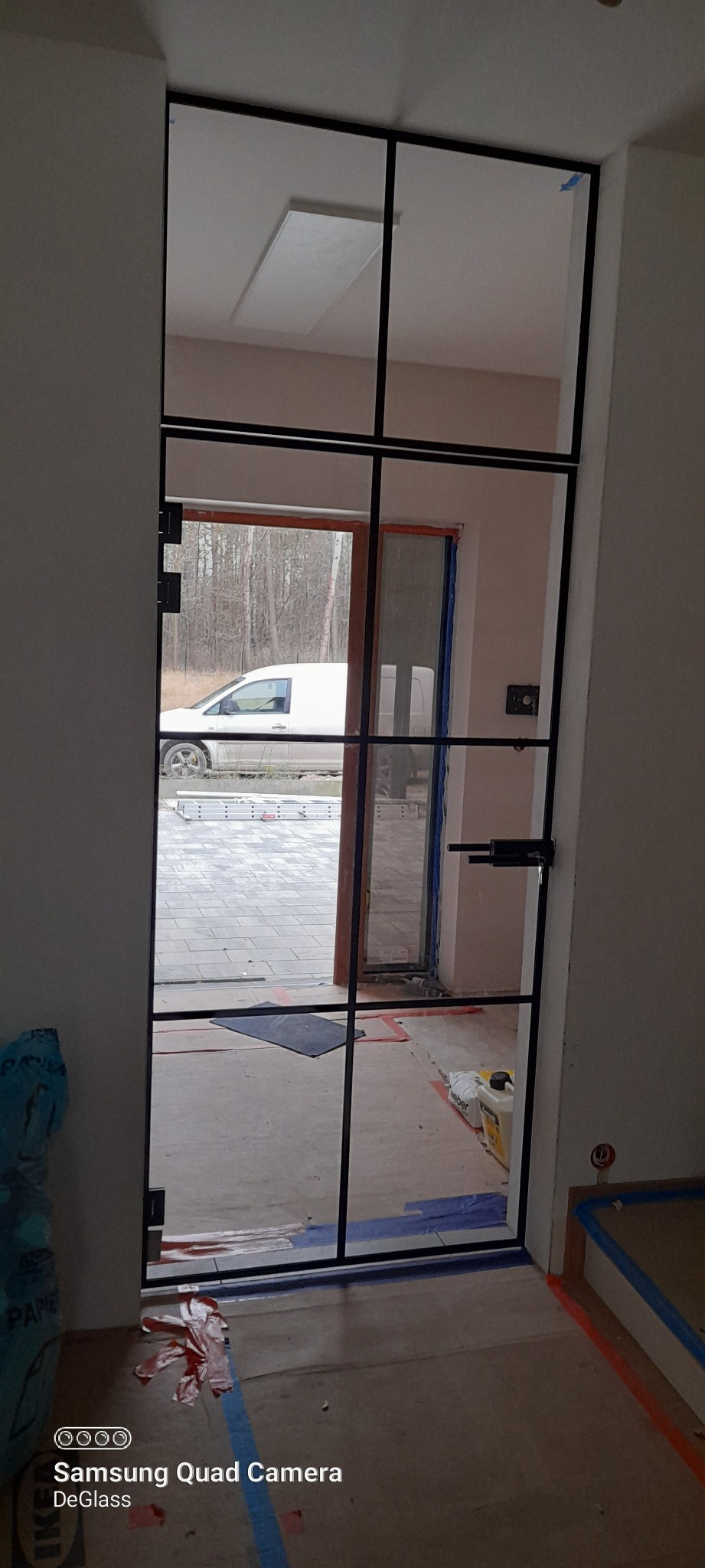 Szklane drzwi w czarnej ramie, widok na zewnątrz z białym samochodem dostawczym i materiały budowlane na podłodze wewnątrz.