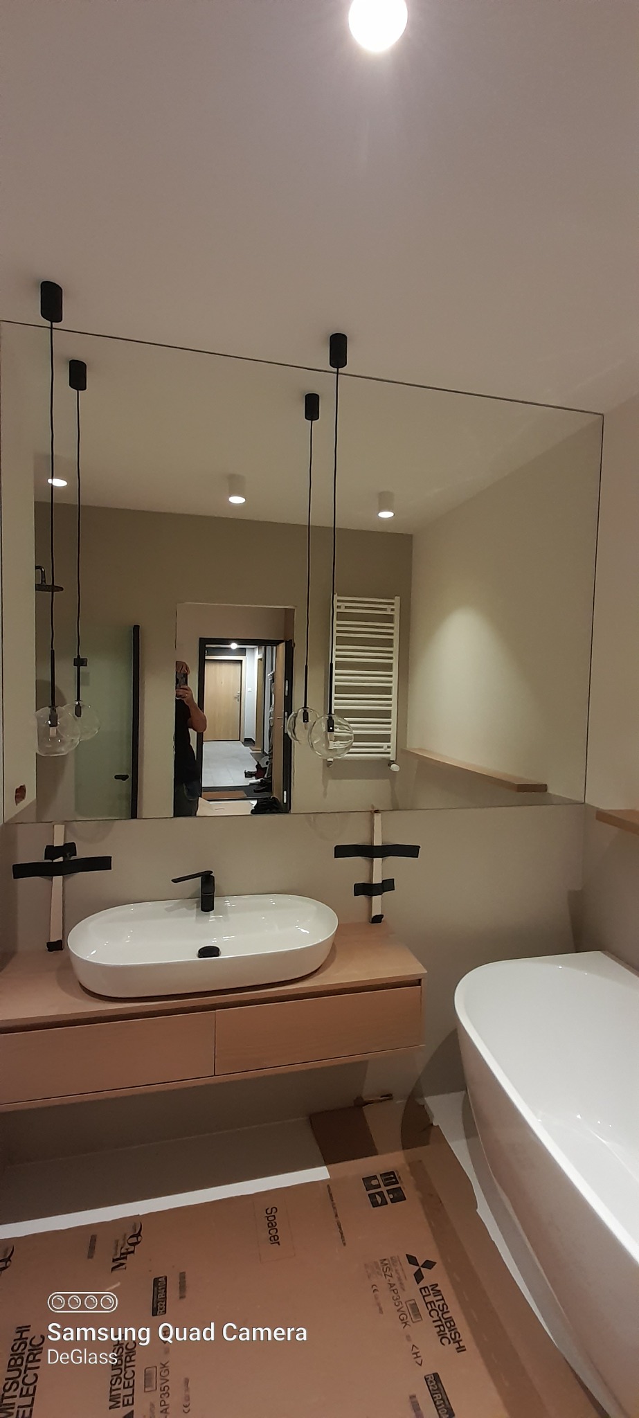 Nowoczesna łazienka z dużym lustrem na ścianie, wiszącą szafką pod umywalką i wiszącymi lampami, widoczne zabezpieczenia lustra taśmą montażową.