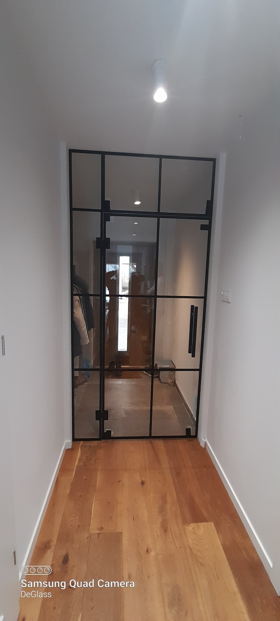 Przeszklone drzwi wewnętrzne z czarną ramą w korytarzu z drewnianą podłogą, widok z perspektywy pomieszczenia.