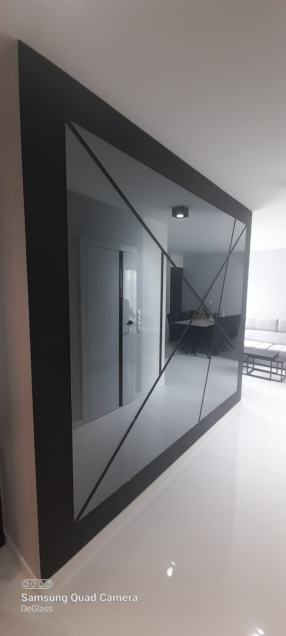 Duże lustro dekoracyjne z czarną ramą i geometrycznymi podziałami, odbijające wnętrze nowoczesnego salonu z białymi ścianami i podłogą.