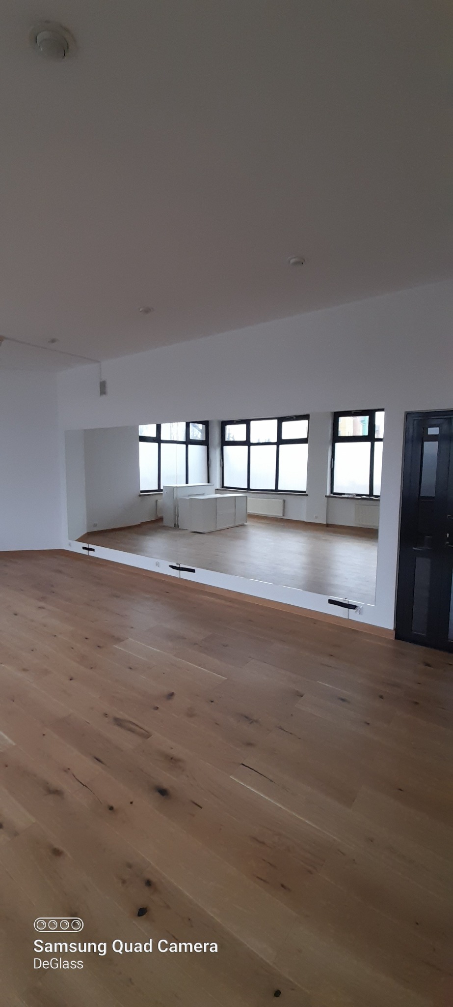 Duże lustro na ścianie w sali tanecznej lub siłowni, zamocowane na wspornikach, odbijające okna z mlecznymi szybami i białe szafki