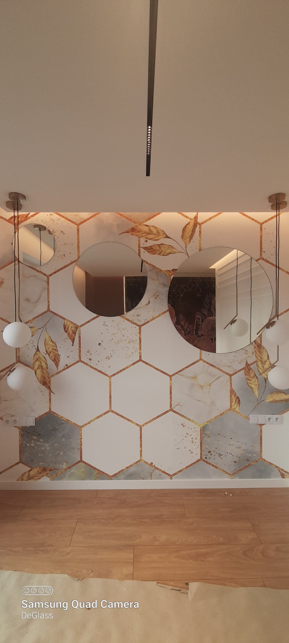 Wnętrze z trzema okrągłymi lustrami bez ram na dekoracyjnej ścianie z motywem plastrów miodu i złotych liści, oświetlone lampami wiszącymi z kulistymi kloszami.