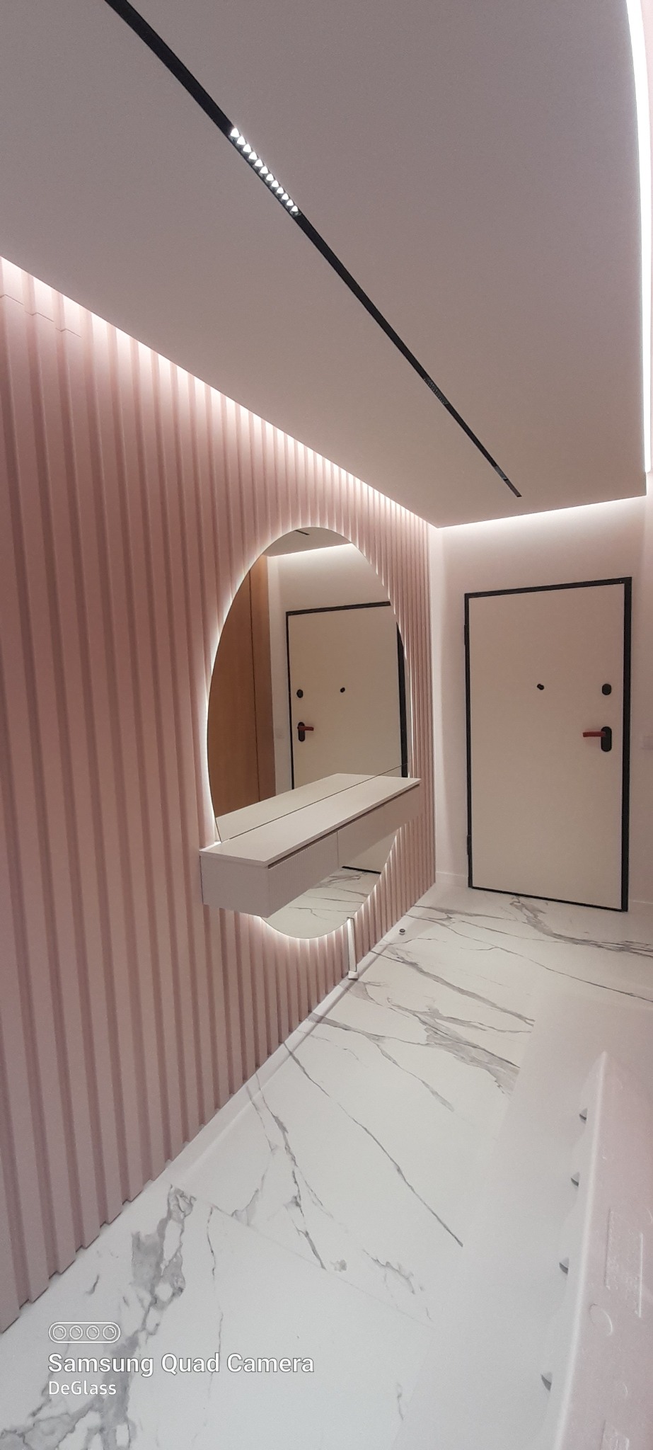 Nowoczesny przedpokój z pudrowo-różową ścianą z pionowymi panelami, okrągłym lustrem z podświetleniem LED i marmurową podłogą.