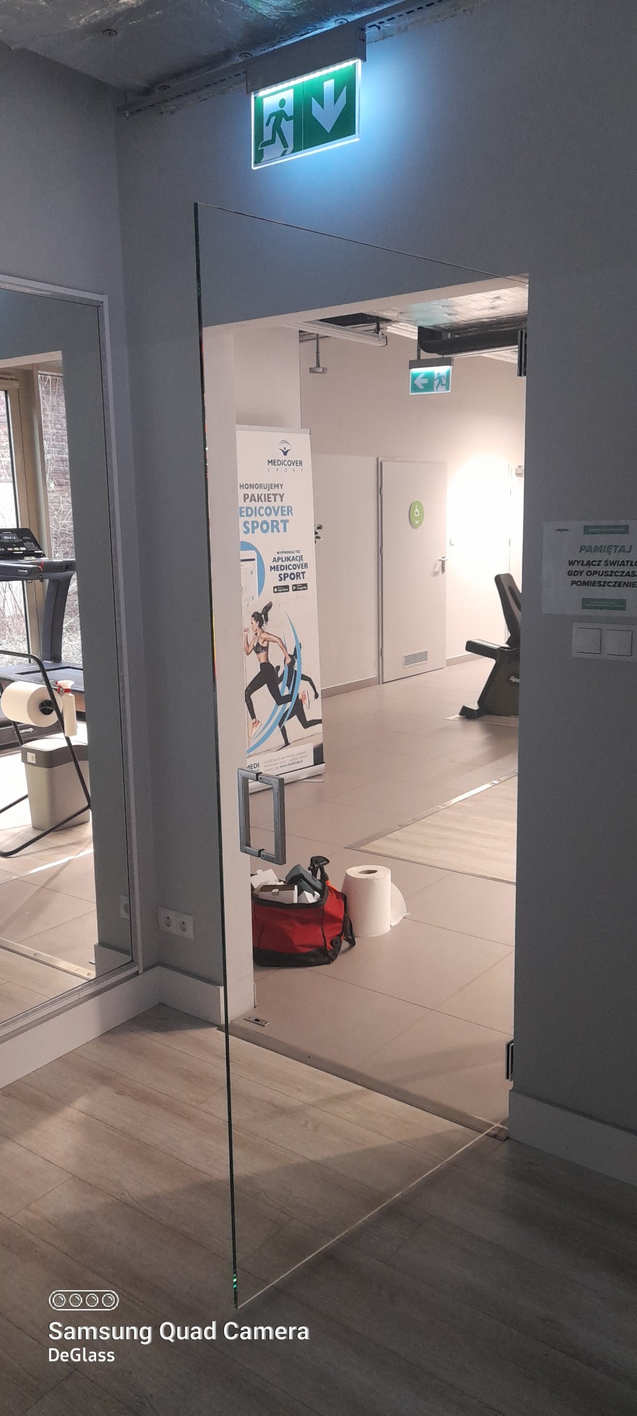 Szklane drzwi w pomieszczeniu, widoczny plakat Medicover Sport i znak ewakuacyjny, podłoga z paneli.