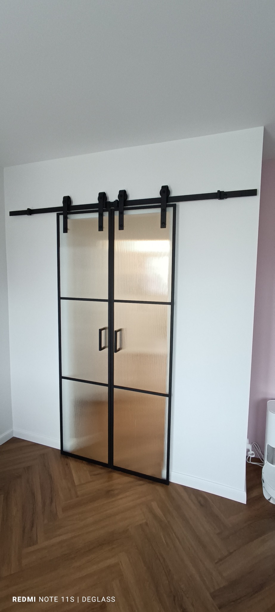 Dwuskrzydłowe przesuwne drzwi szklane z czarną ramą i poziomymi podziałami, zamontowane na szynie w jasnym wnętrzu z parkietem w jodełkę.