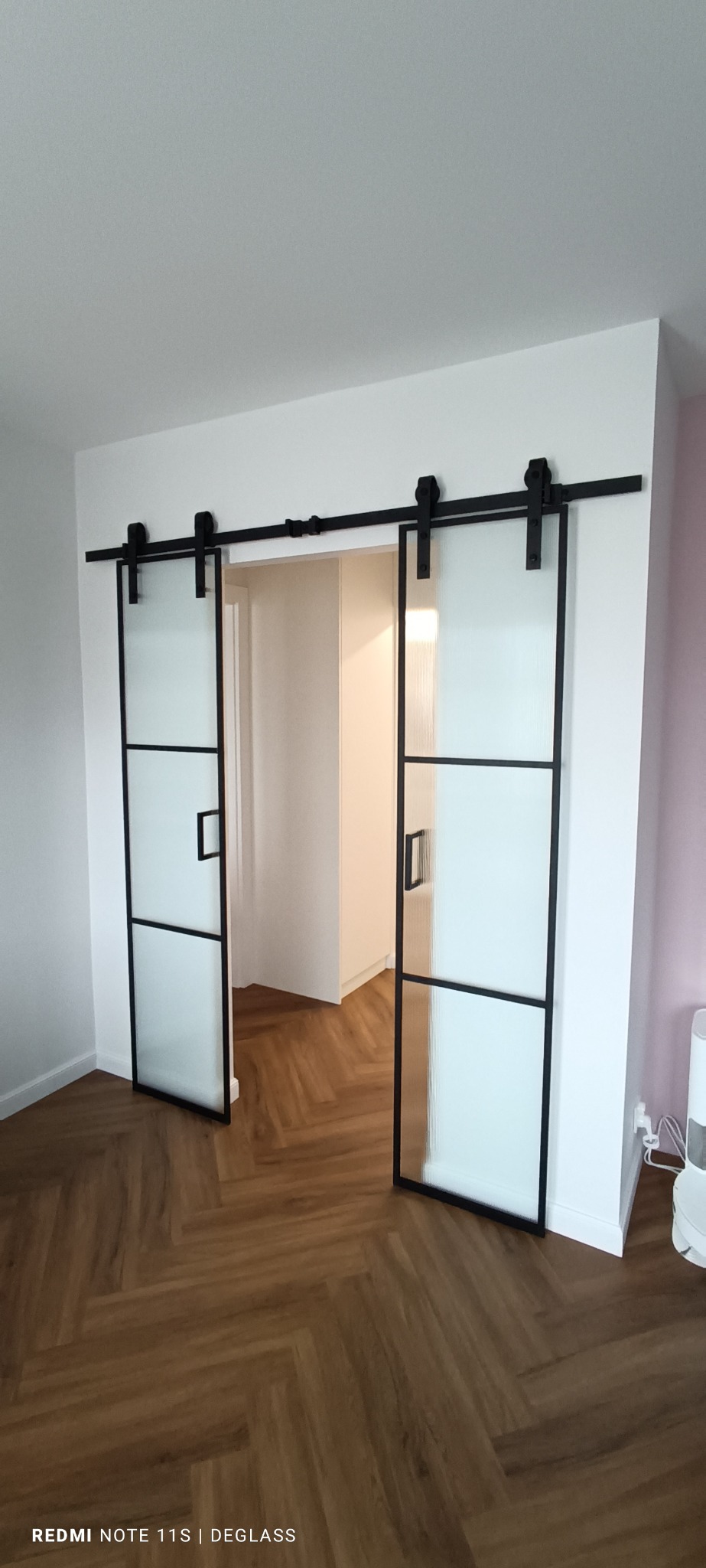 Nowoczesne drzwi przesuwne z matowego szkła w czarnej ramie, zamontowane na szynie, widok na wnętrze z parkietem ułożonym w jodełkę.