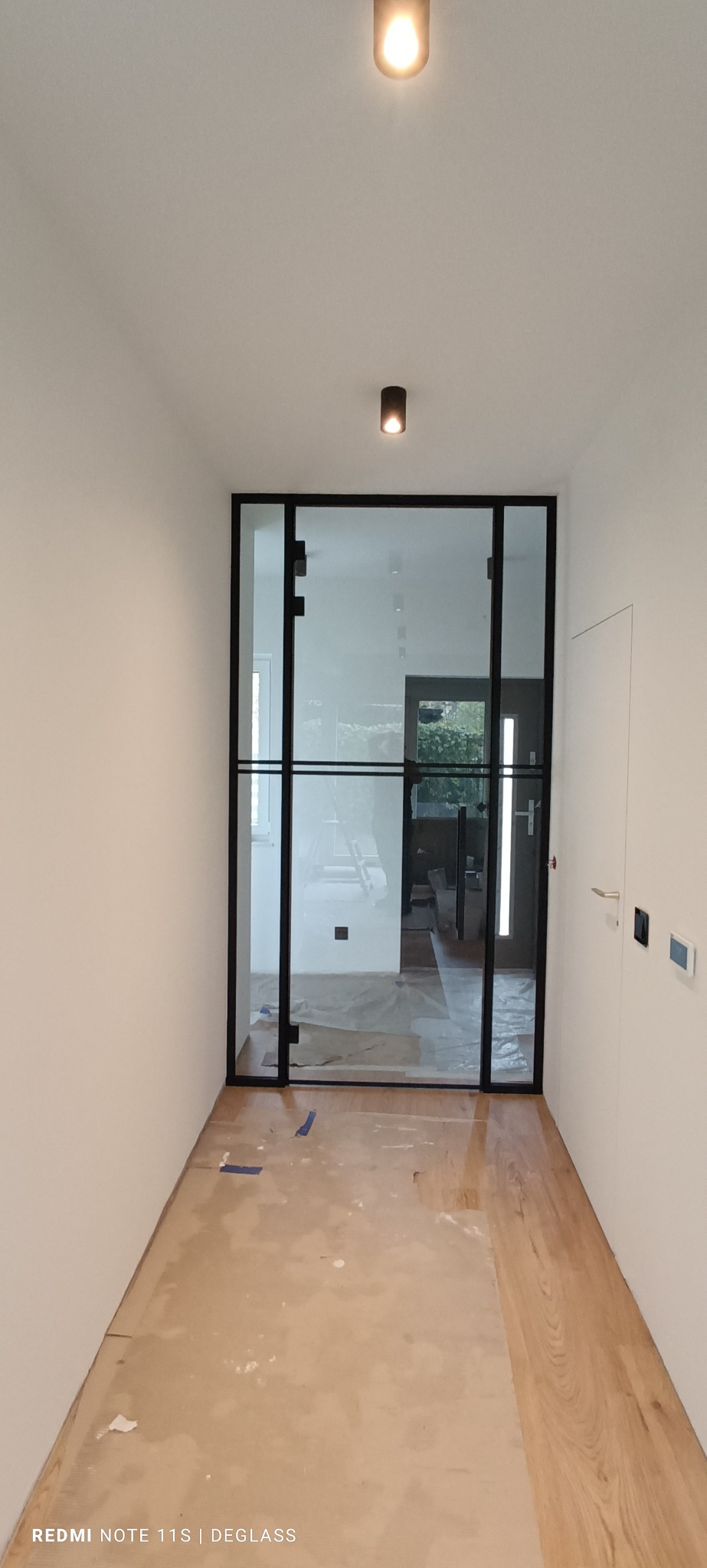 Szklane drzwi w czarnej ramie, oddzielające korytarz w mieszkaniu w Warszawie, widok na sąsiednie pomieszczenie przez szybę, podłoga zabezpieczona folią malarską.
