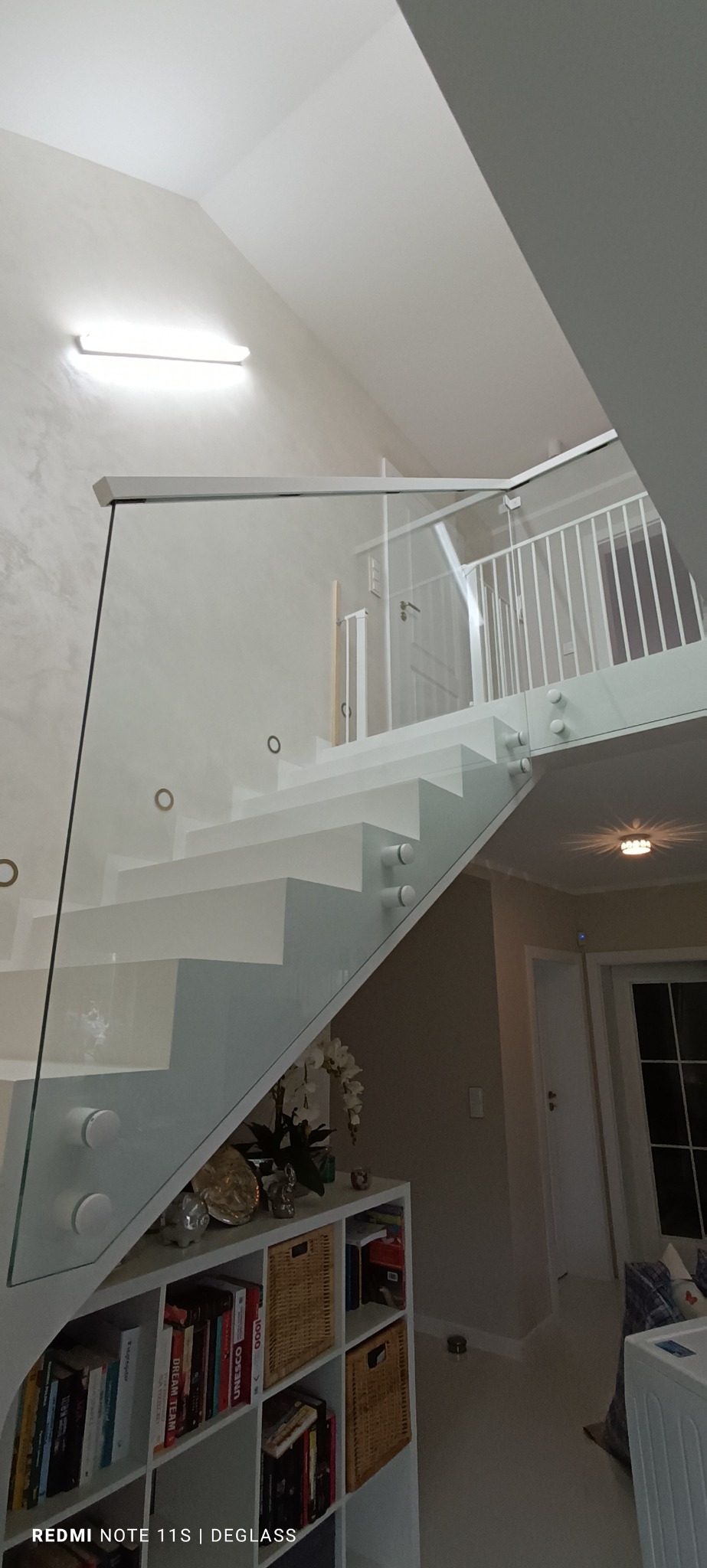 Nowoczesne, białe schody z szklaną balustradą mocowaną punktowo, widok z dołu na górę, z elementami dekoracyjnymi w tle.