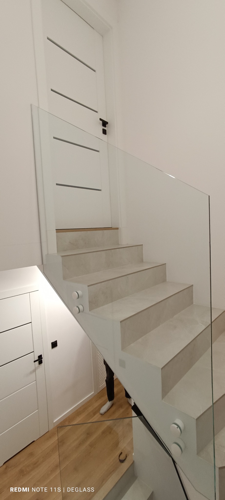 Nowoczesne schody z jasnymi stopniami i szklaną balustradą mocowaną na okrągłych uchwytach, widok z dołu na białe drzwi z poziomymi frezowaniami.