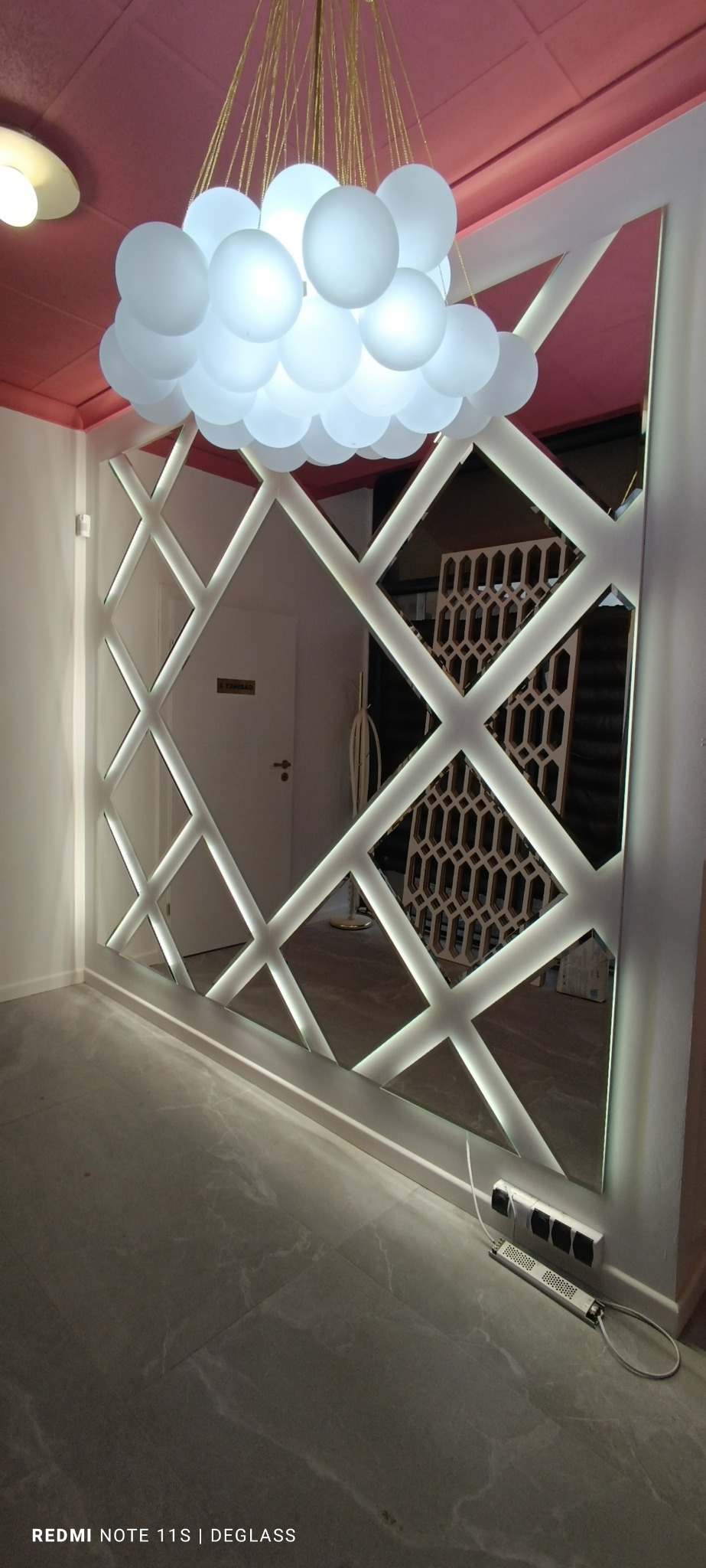 Ściana z dużym lustrem w romby z podświetleniem LED, odbijająca kratownicową dekorację i białe drzwi, oświetlona lampą w kształcie chmury z kul.