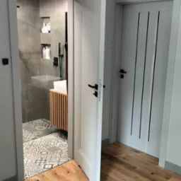 Widok na wykończoną łazienkę z kabiną prysznicową typu walk-in, umywalką nablatową na szafce z lamelami i wzorzystą podłogą, widziany przez otwarte białe drzwi z czarną klamką, na pierwszym planie...