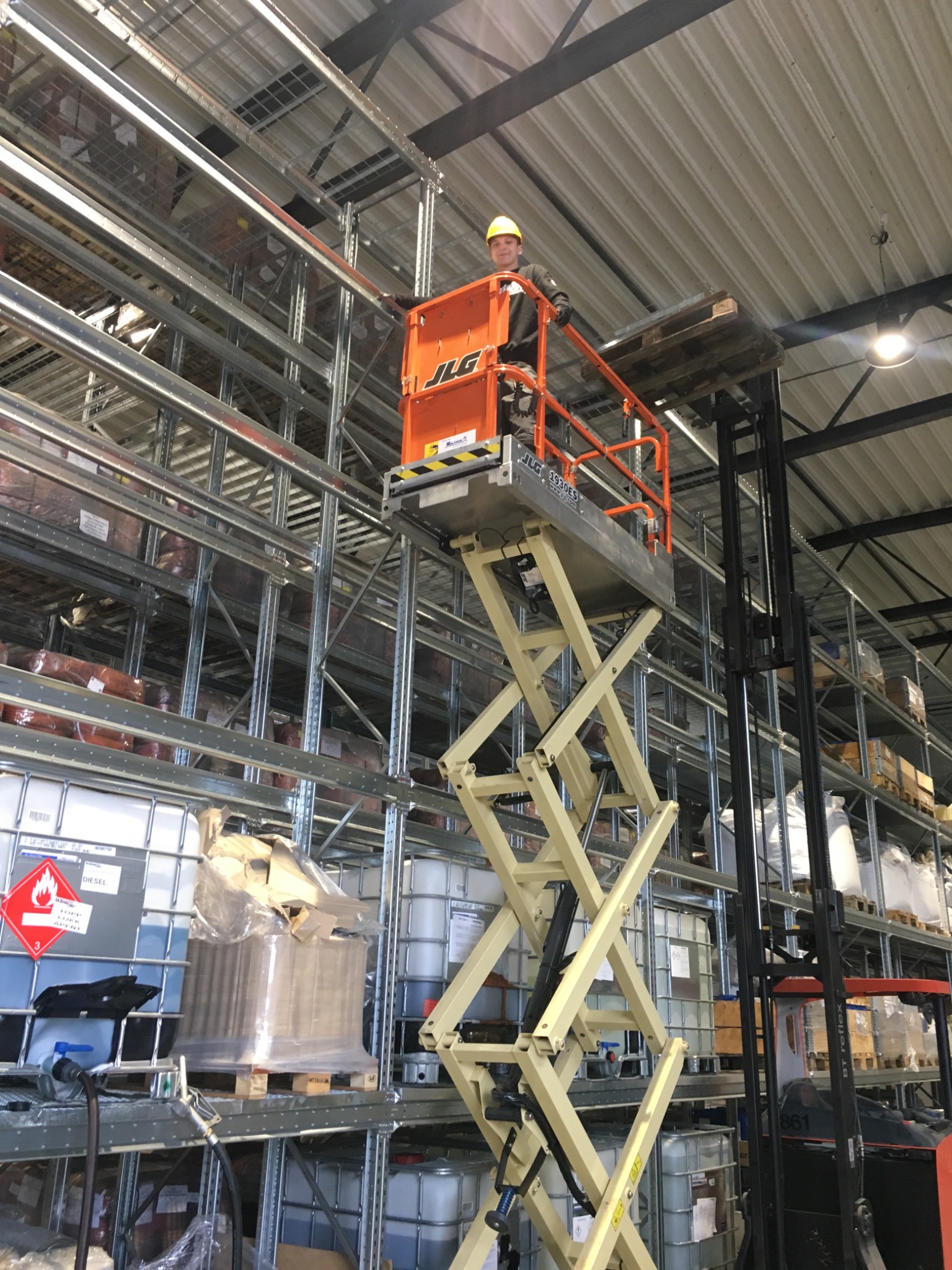 Pracownik w kasku na podnośniku nożycowym JLG, umieszczający paletę na wysokim regale magazynowym z różnymi towarami, widok z dołu w hali magazynowej.