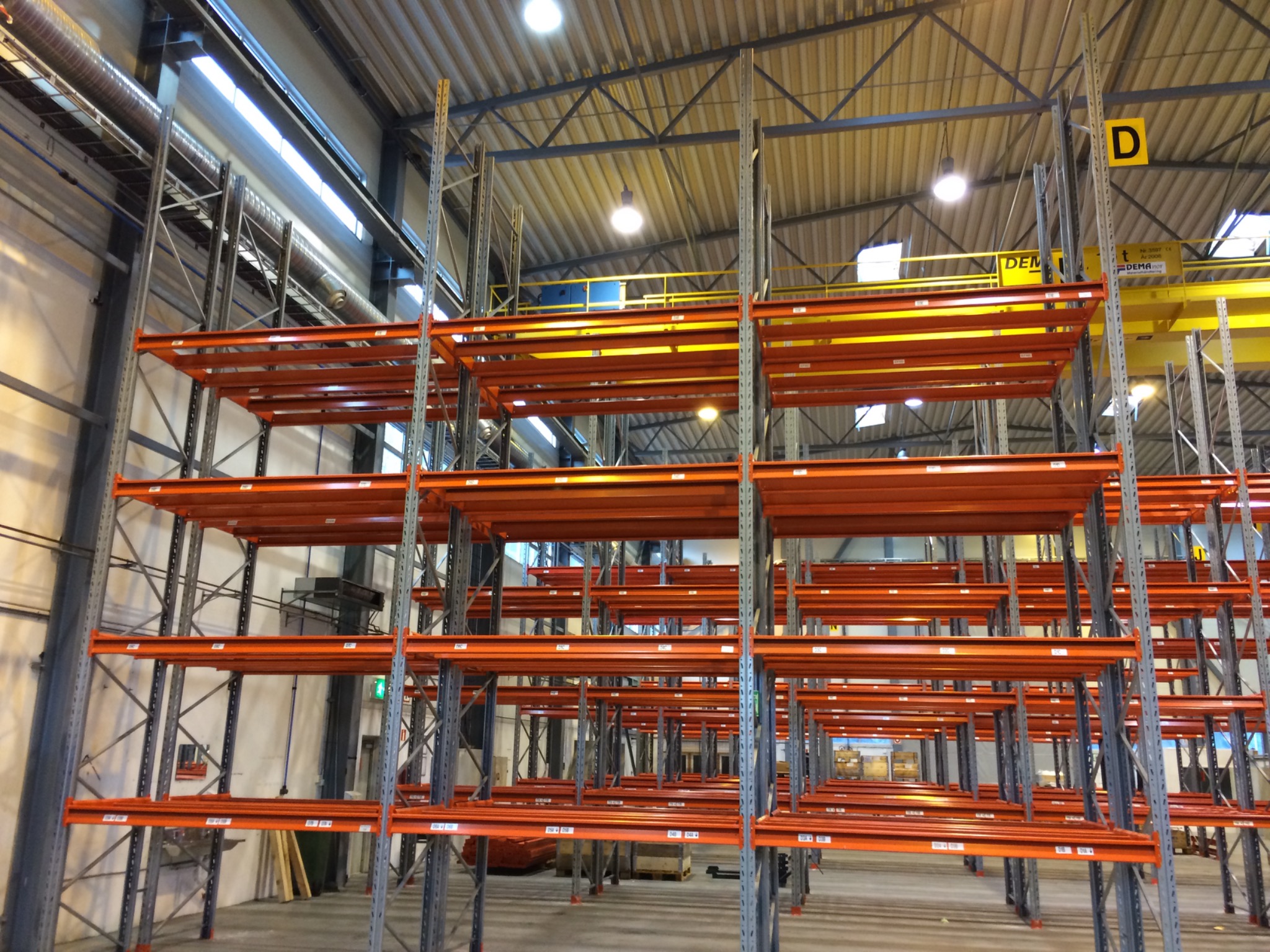 Puste, pomarańczowe regały magazynowe z szarymi słupkami, widok z dołu, z widoczną konstrukcją dachu magazynu i oświetleniem.