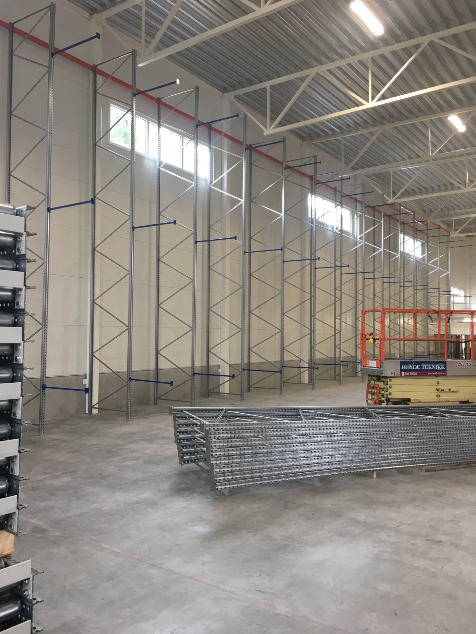 Wysokie, metalowe regały magazynowe z niebieskimi wspornikami, częściowo zamontowane w hali magazynowej z oknami i podnośnikiem koszowym w tle, oraz stosy elementów do montażu na podłodze.