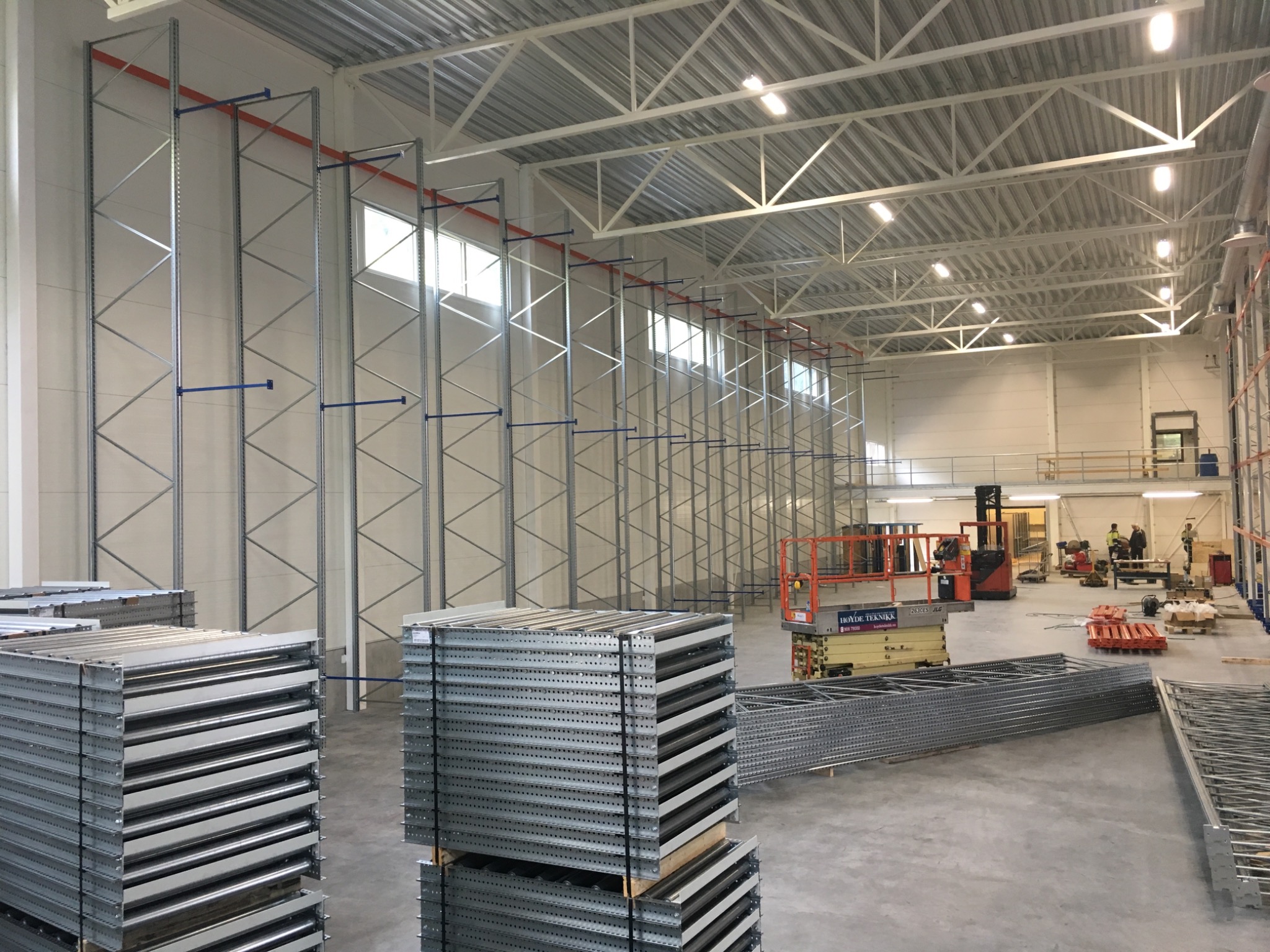 Wnętrze hali magazynowej z zamontowanymi pionowymi elementami konstrukcji regałów, palety z metalowymi półkami i podnośnik nożycowy w tle.