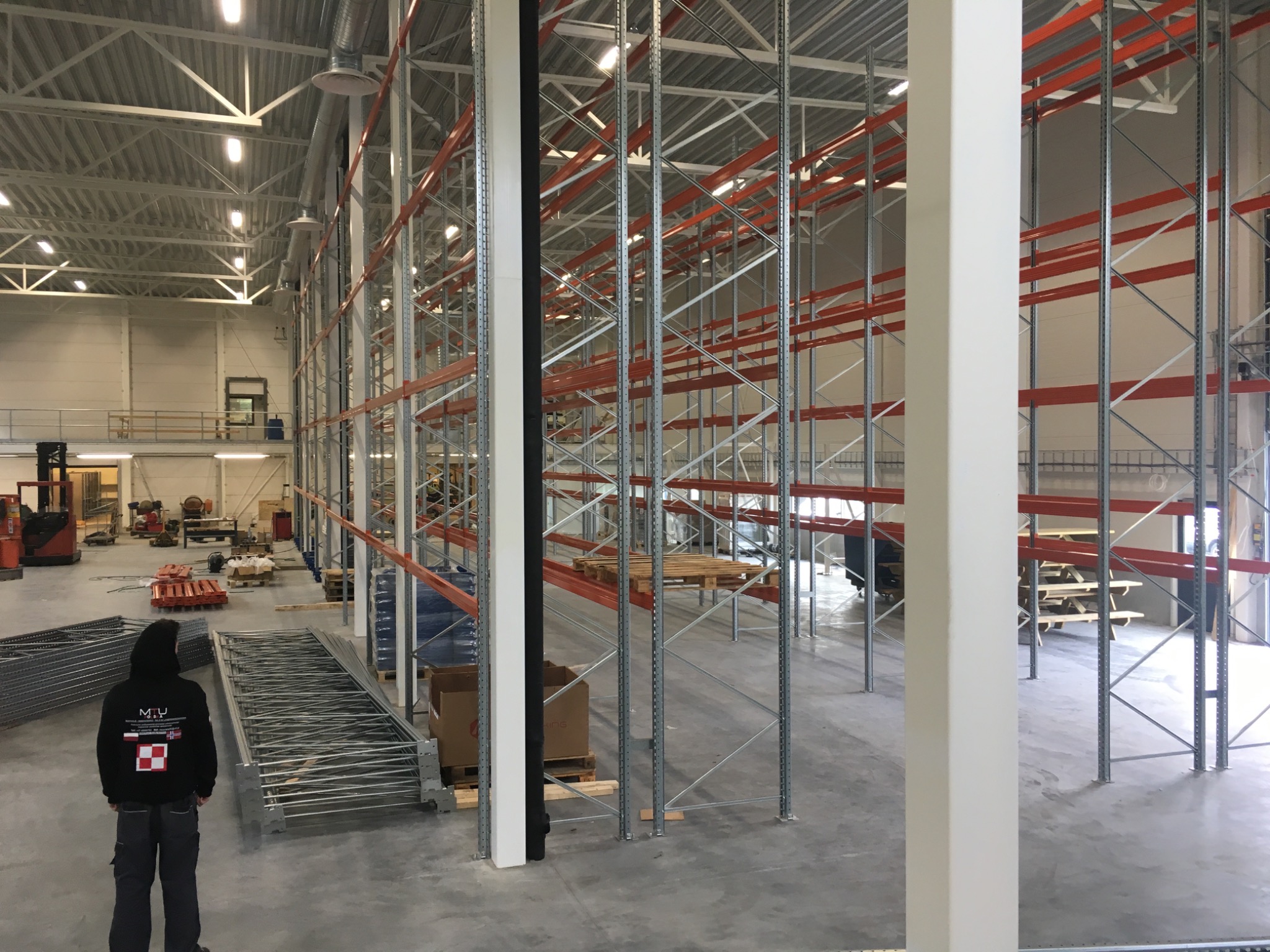 Wnętrze magazynu z zamontowanymi, metalowymi regałami z pomarańczowymi belkami, widoczny pracownik w czarnej bluzie z logo firmy, oraz wózek widłowy w tle.