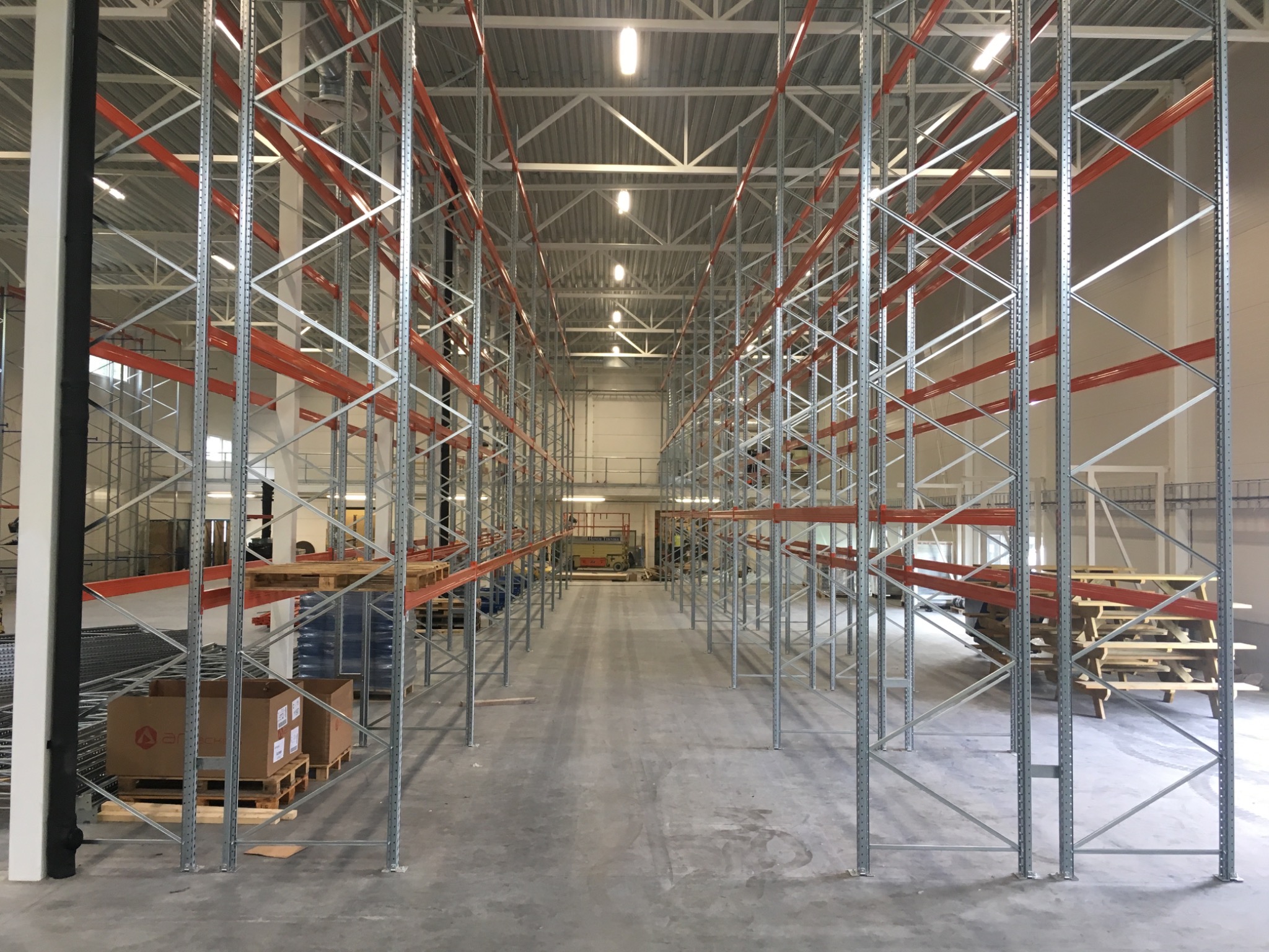 Długa perspektywa rzędów metalowych regałów z czerwonymi poprzeczkami w przestronnym, jasnym magazynie, z paletami i kartonami u podstawy.