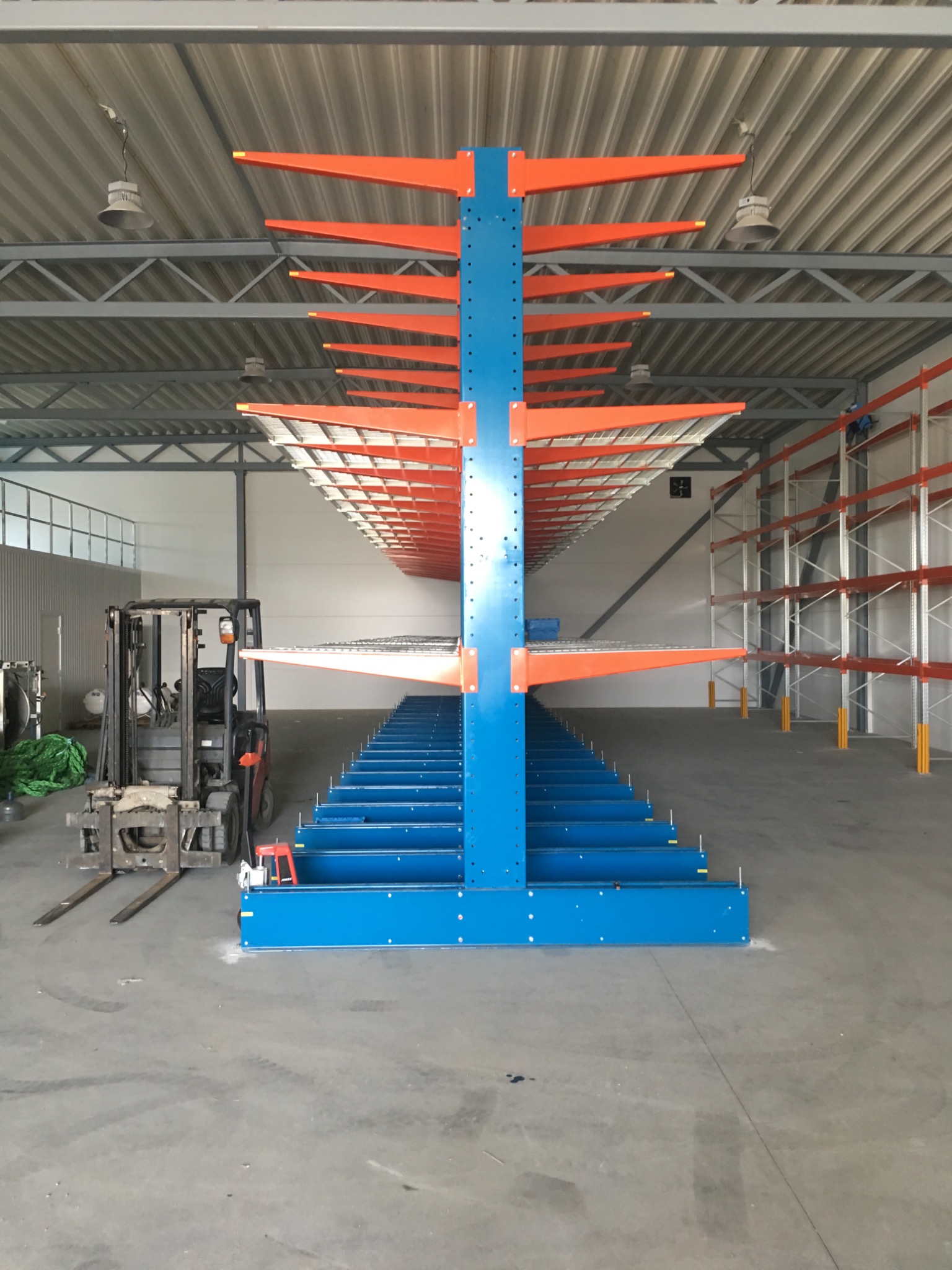 Niebiesko-pomarańczowy regał magazynowy wspornikowy z widocznym wózkiem widłowym w tle, oświetlony lampami przemysłowymi, w przestronnym magazynie.