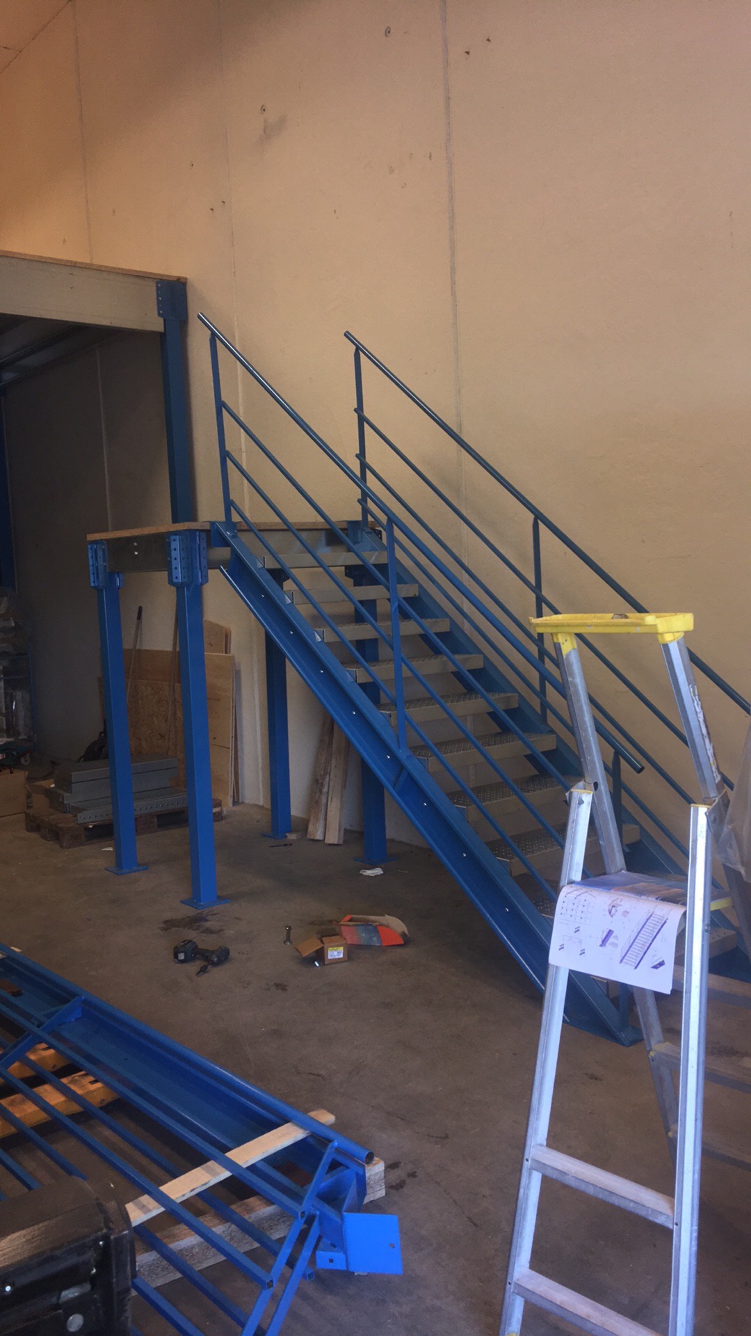 Niebieska metalowa antresola schodowa w trakcie montażu w hali, widoczna drabina z instrukcją montażu, elementy konstrukcyjne na podłodze.
